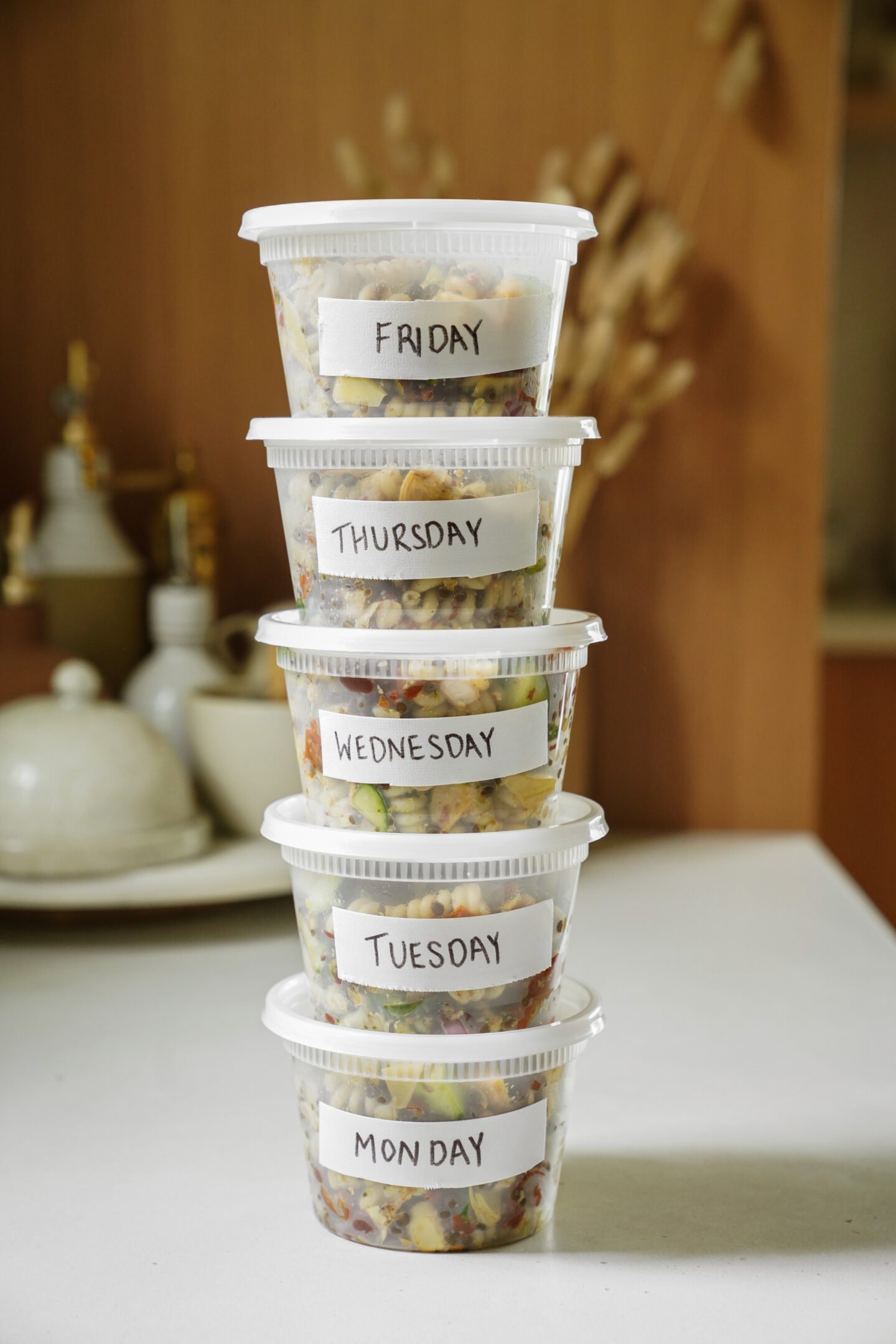Five clear food containers stacked on a kitchen counter, each labeled with a day of the week from Monday to Friday, containing pre-prepared meals.