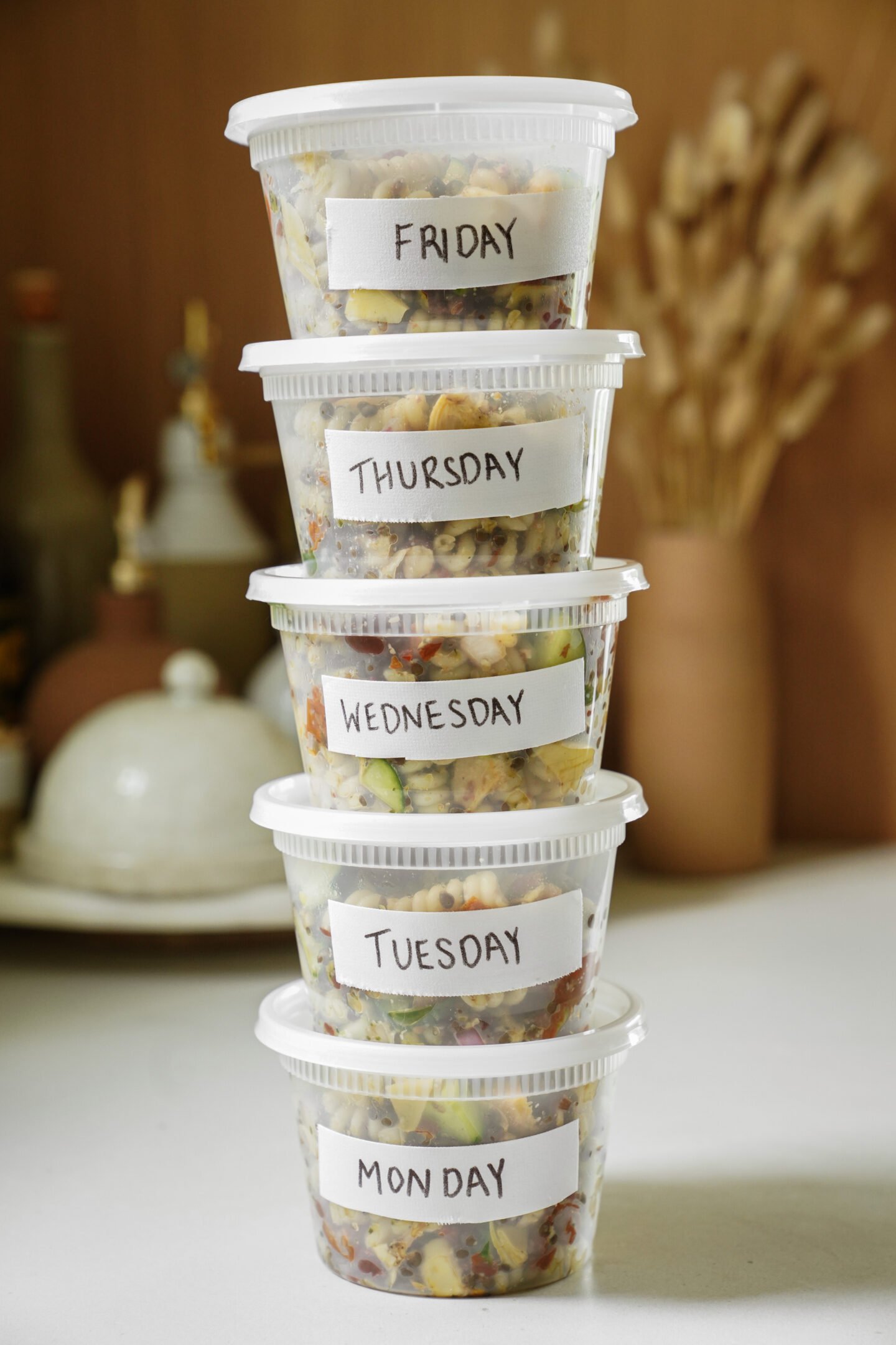 Five plastic food containers are stacked, each labeled with a weekday from Monday to Friday, filled with colorful chopped salads or meal prep ingredients, set on a kitchen counter with blurred background objects.