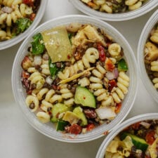 A close-up of a plastic container filled with pasta salad containing spiral pasta, lentils, chopped cucumber, artichoke hearts, red onion, olives, and herbs, surrounded by other similar containers.