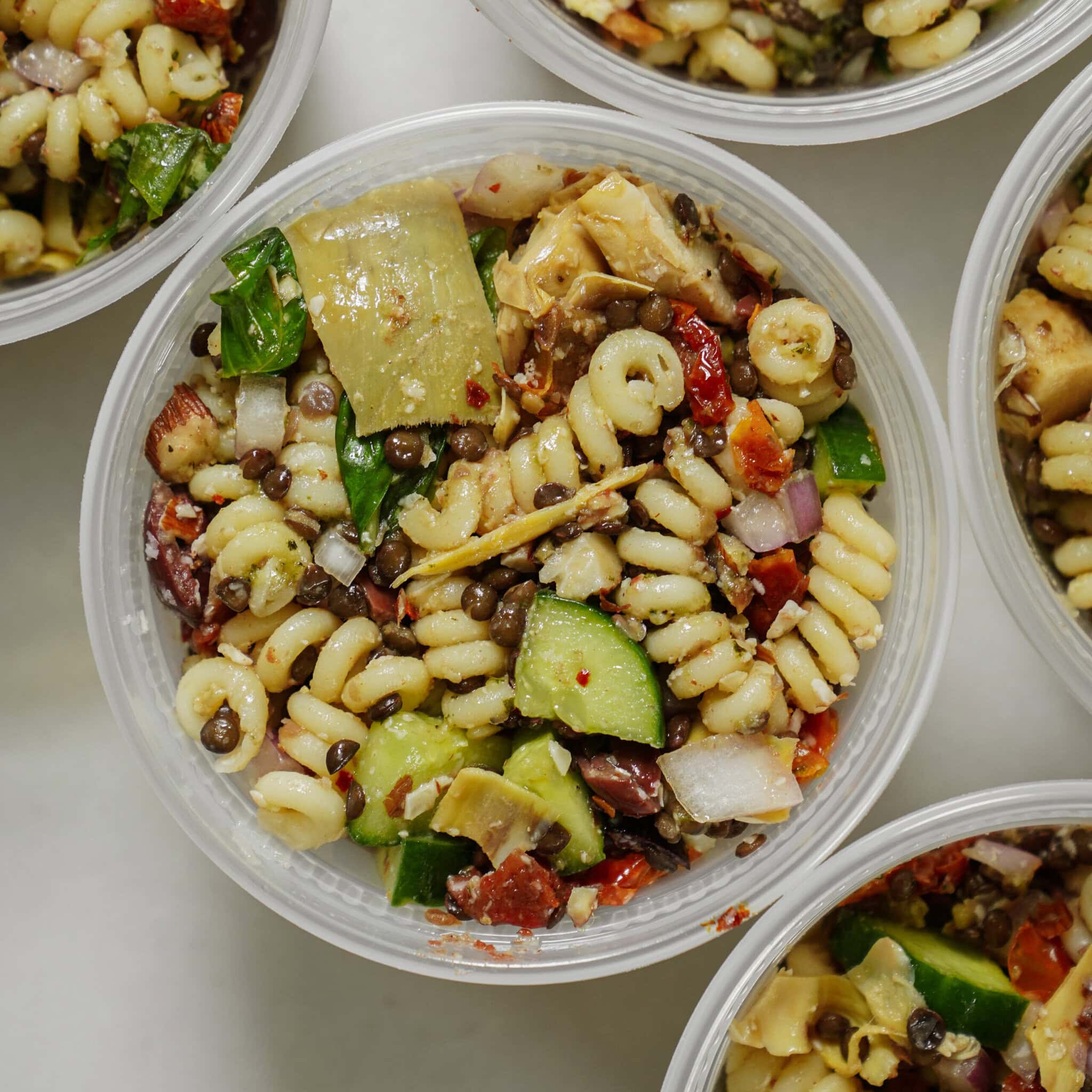 A close-up of a plastic container filled with pasta salad containing spiral pasta, lentils, chopped cucumber, artichoke hearts, red onion, olives, and herbs, surrounded by other similar containers.