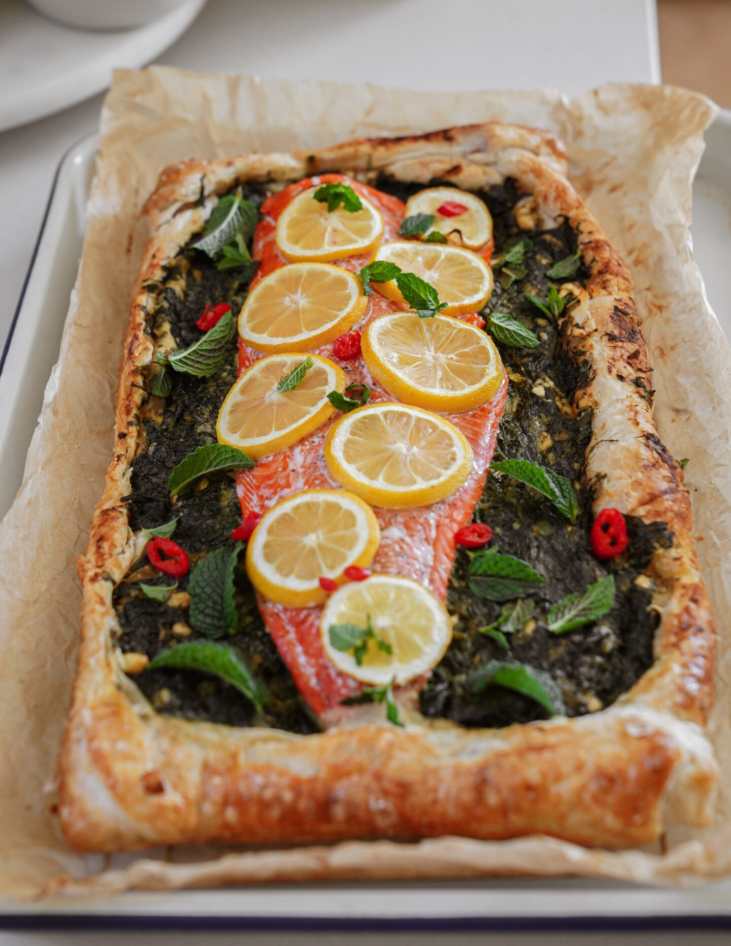 A rectangular puff pastry tart topped with a fillet of salmon, lemon slices, fresh mint leaves, red chili slices, and a green herb spread, baked on parchment paper.
