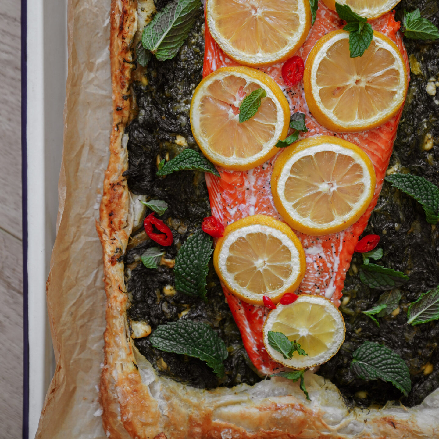 A baked salmon fillet topped with lemon slices and herbs sits on a puff pastry crust, garnished with fresh mint leaves and red chili slices. The pastry edges are golden brown.