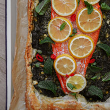 A baked salmon fillet topped with lemon slices and herbs sits on a puff pastry crust, garnished with fresh mint leaves and red chili slices. The pastry edges are golden brown.