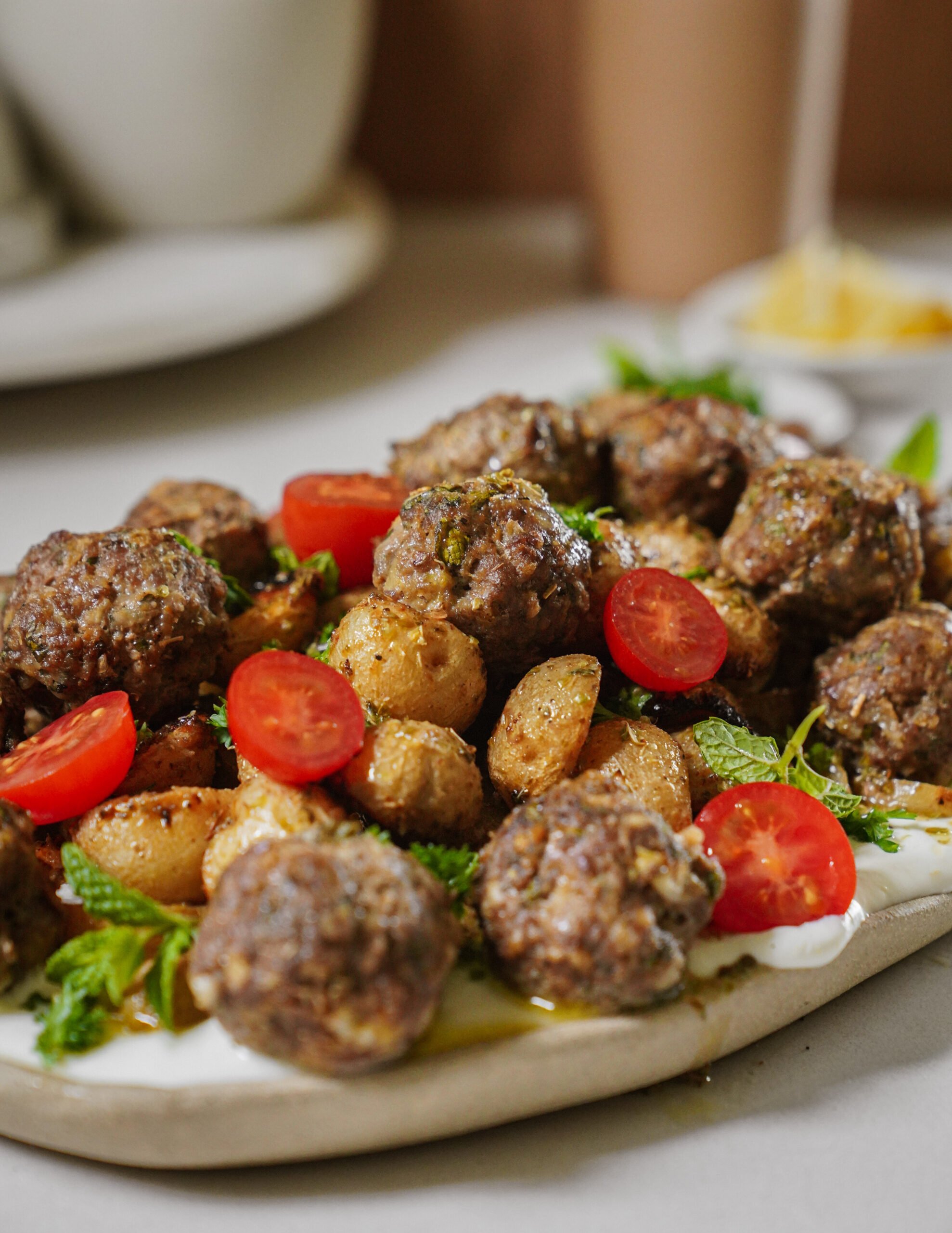 A plate of meatballs served with roasted potatoes, halved cherry tomatoes, fresh mint, and a creamy white sauce on a white platter.