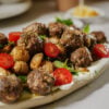 A plate of meatballs served on a bed of creamy sauce, garnished with roasted baby potatoes, fresh mint leaves, and sliced cherry tomatoes.