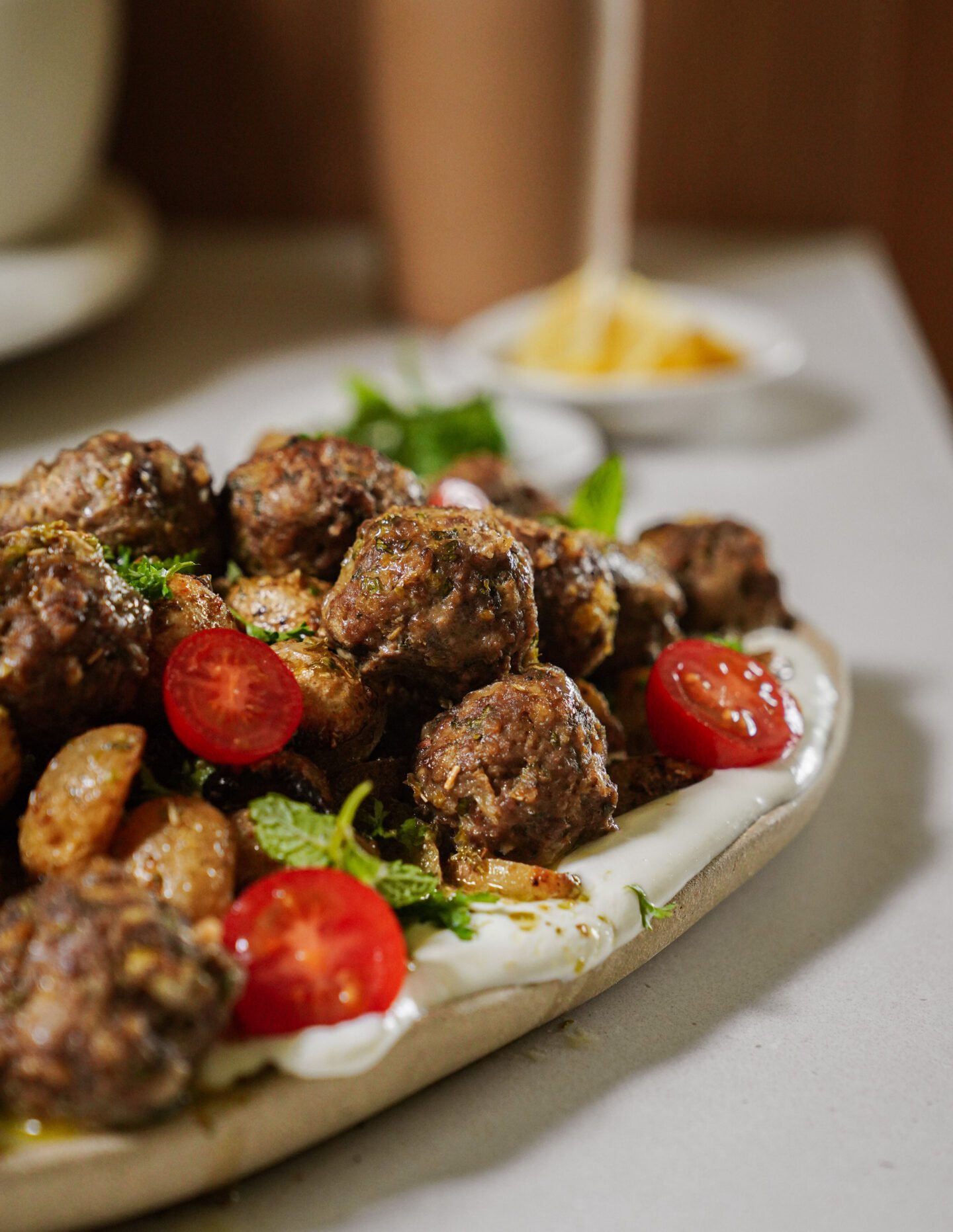 A ceramic plate with meatballs, sliced cherry tomatoes, roasted potatoes, fresh herbs, and a creamy white sauce, placed on a light surface. A small bowl of grated cheese is blurred in the background.