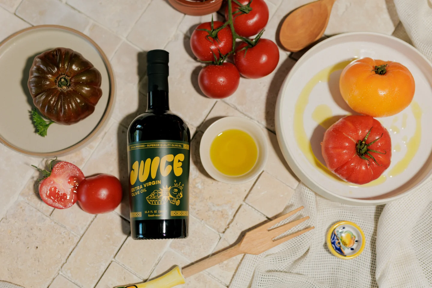 A bottle of extra virgin olive oil labeled JUICE is on a tiled surface, surrounded by various tomatoes, a small bowl of olive oil, a wooden fork, a small dish, and a wooden spoon.