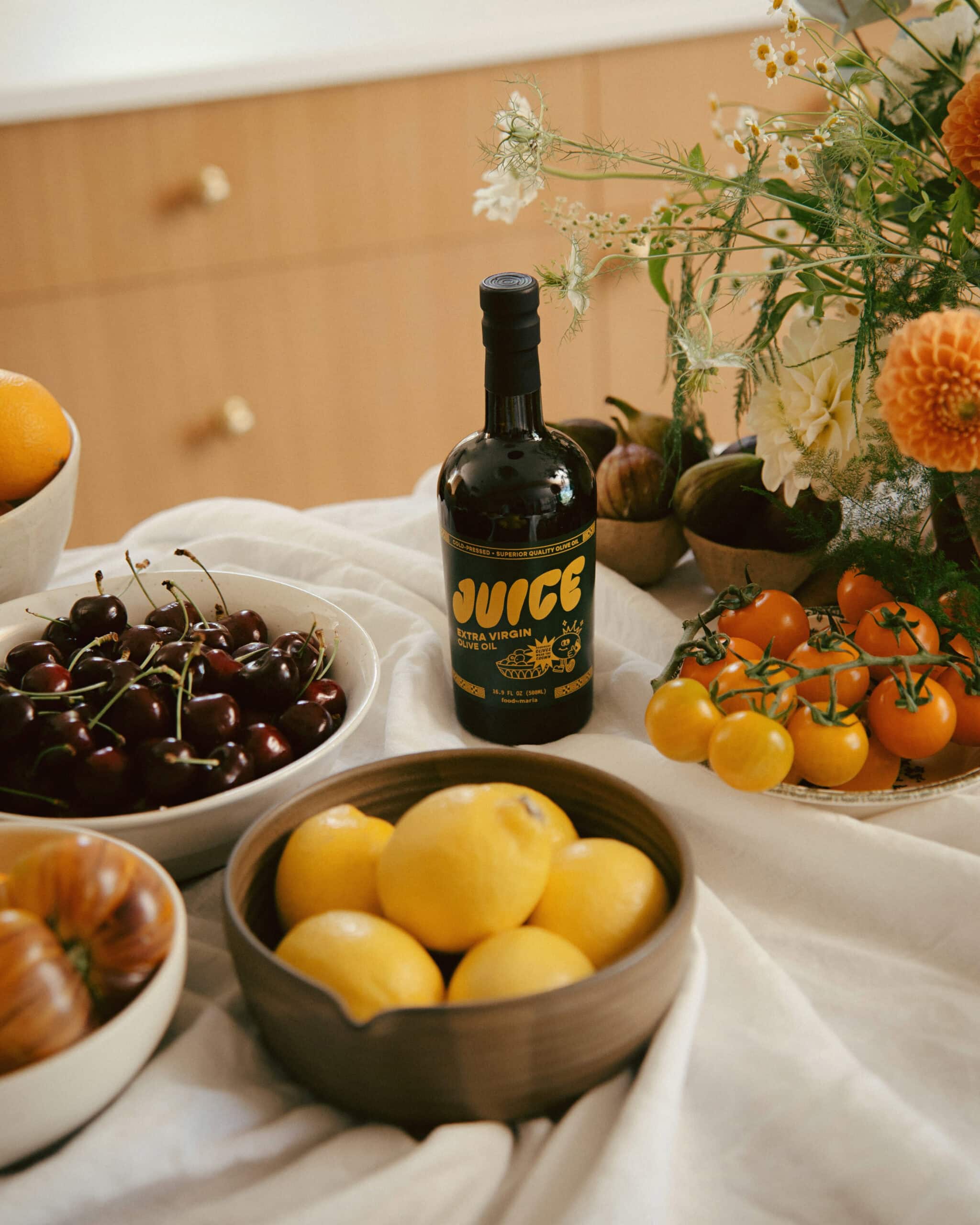 A bottle of olive oil labeled “JUICE” sits on a table with bowls of lemons, cherries, and heirloom tomatoes, alongside yellow cherry tomatoes and a bouquet of orange and white flowers.