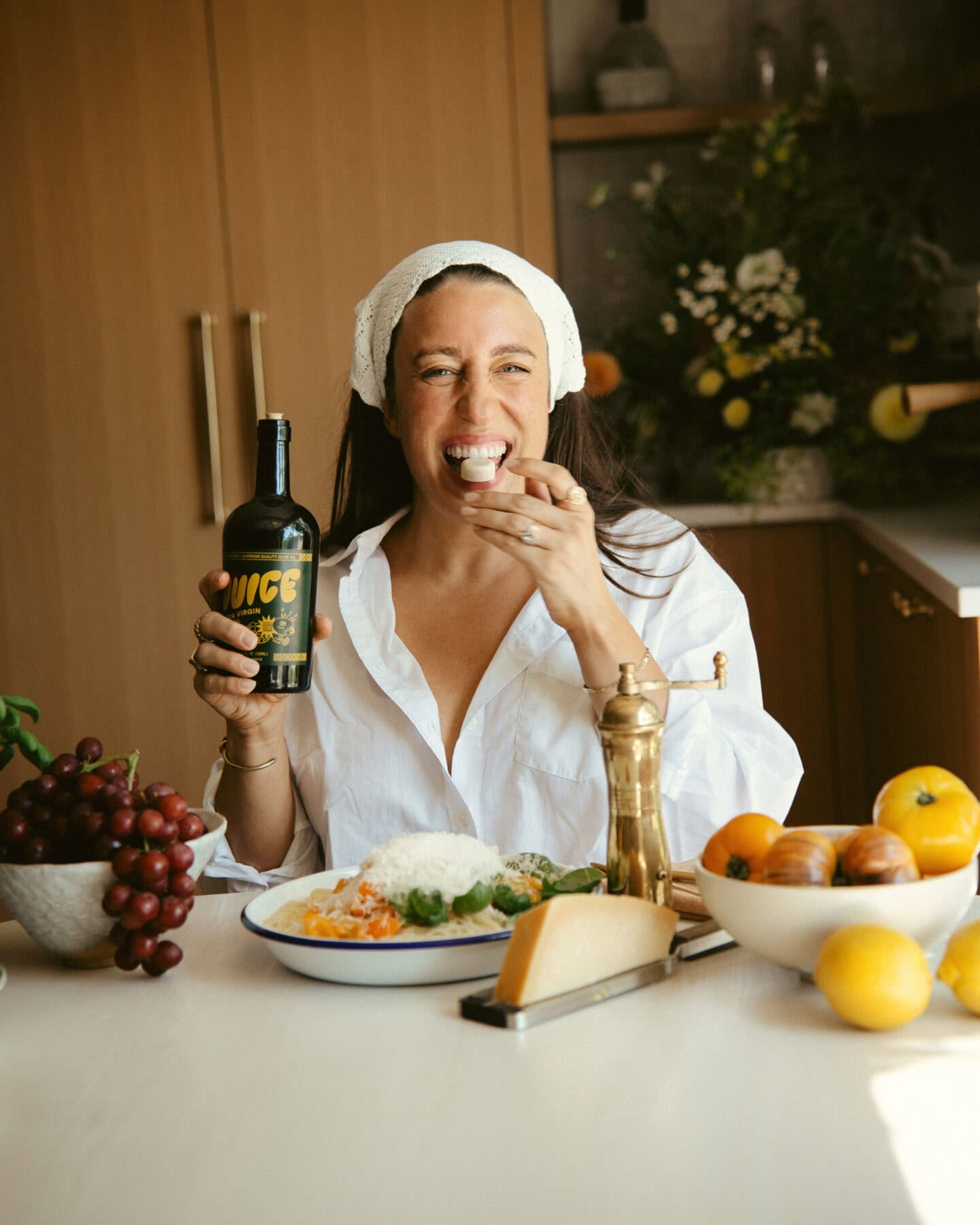 A woman wearing a white shirt and headscarf sits at a table with a plate of food, holding a bottle labeled Juice in one hand and eating with the other, smiling. The table has fruit, cheese, and kitchen items.