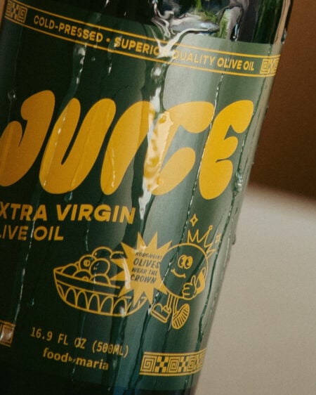 A close-up of a green bottle labeled JUICE EXTRA VIRGIN OLIVE OIL in bold yellow letters, with a cartoon olive character and a small bowl of olives illustrated on the label.