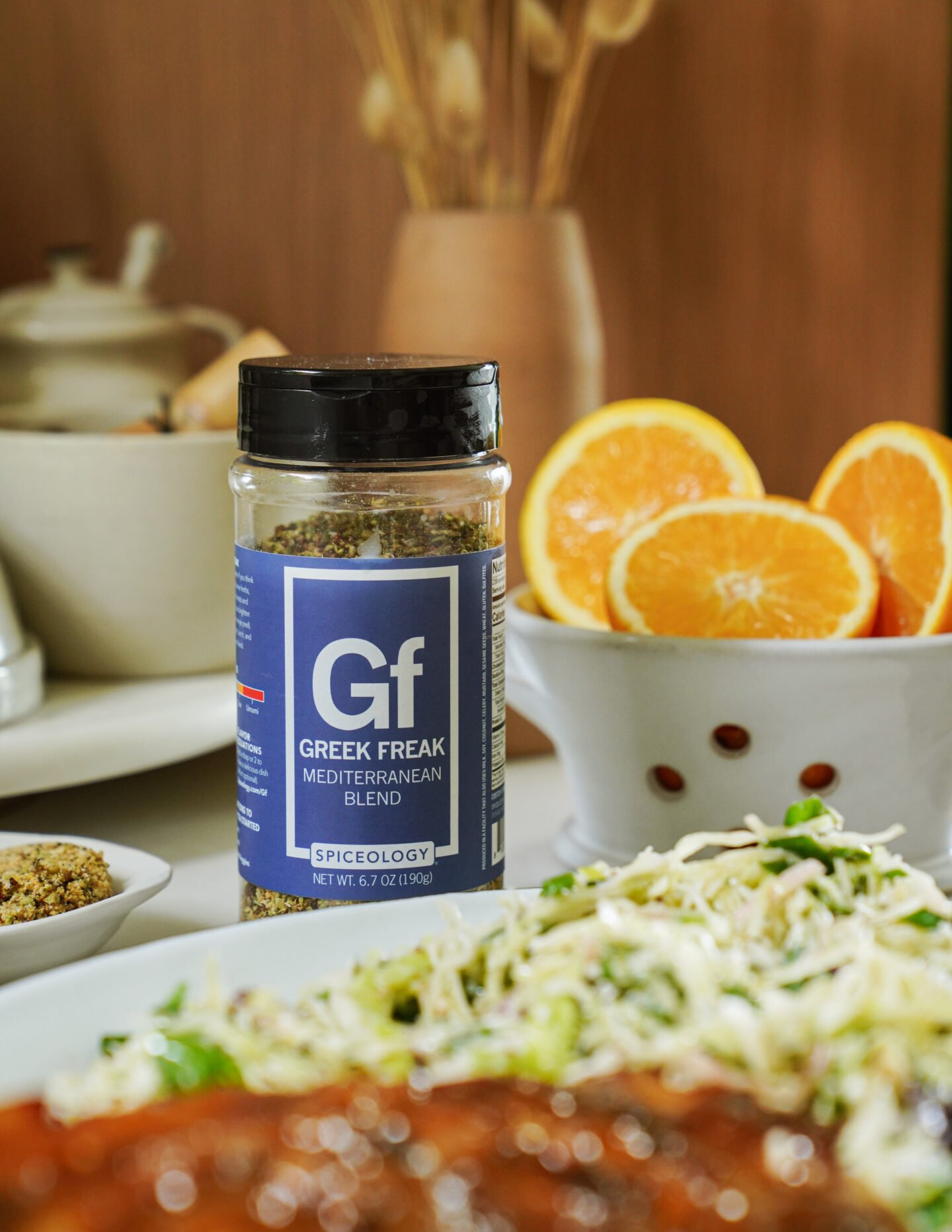 A jar of Greek Freak Mediterranean Blend seasoning sits on a table beside a bowl of sliced oranges and a plate of food, with blurred kitchen items in the background.