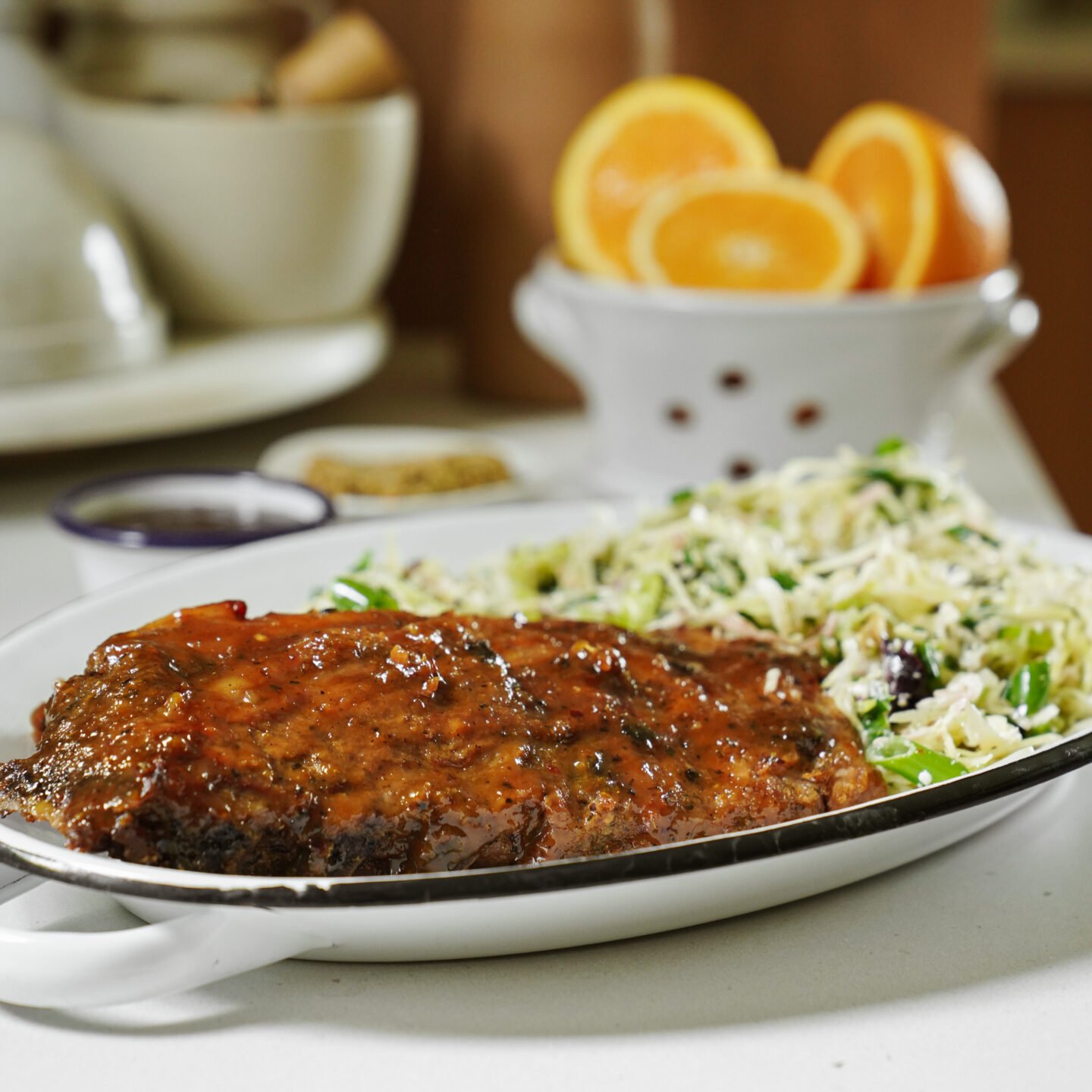 A plate with glazed grilled meat and a side of shredded salad sits on a table; halved oranges and a white bowl are blurred in the background.