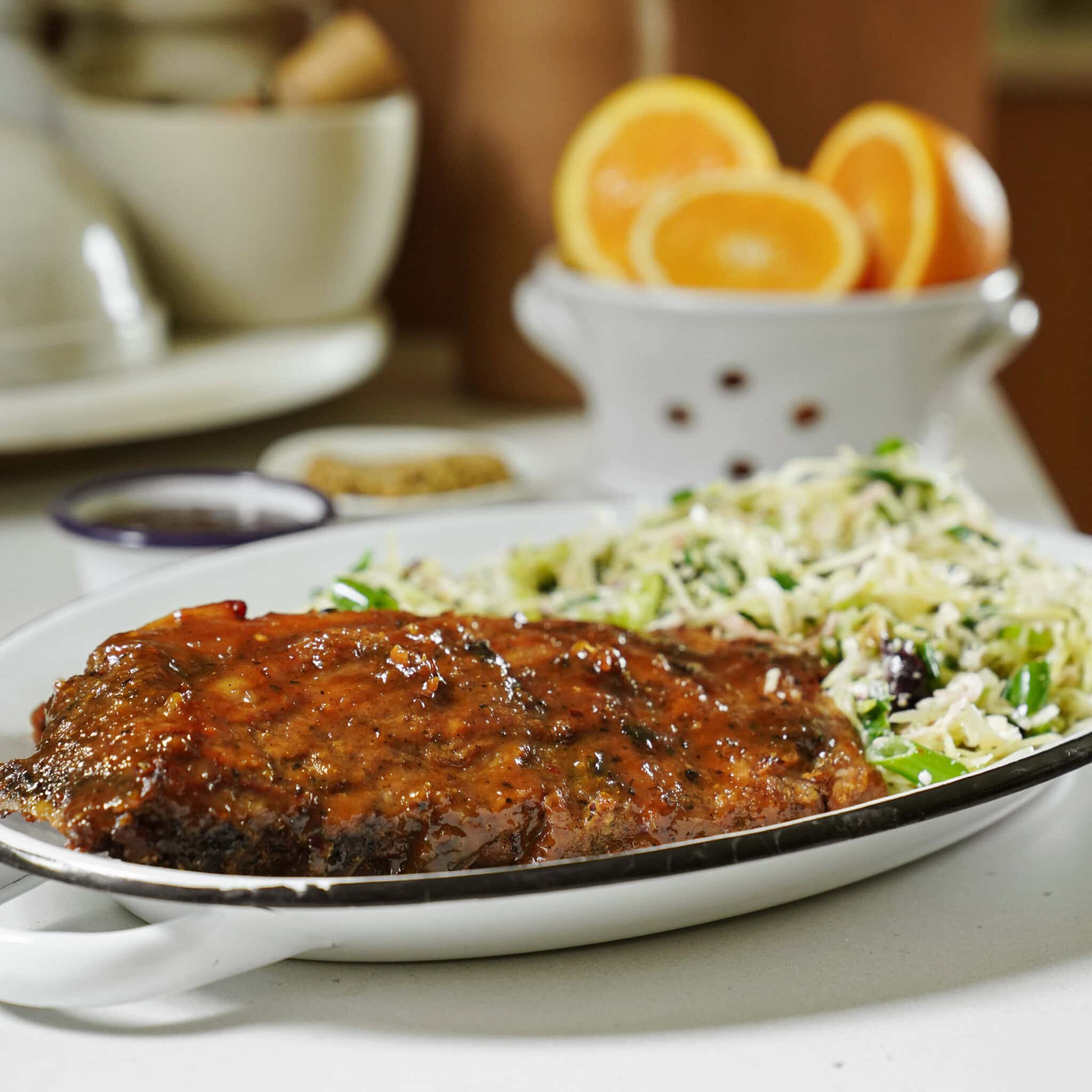 A plate with glazed grilled meat and a side of shredded salad sits on a table; halved oranges and a white bowl are blurred in the background.