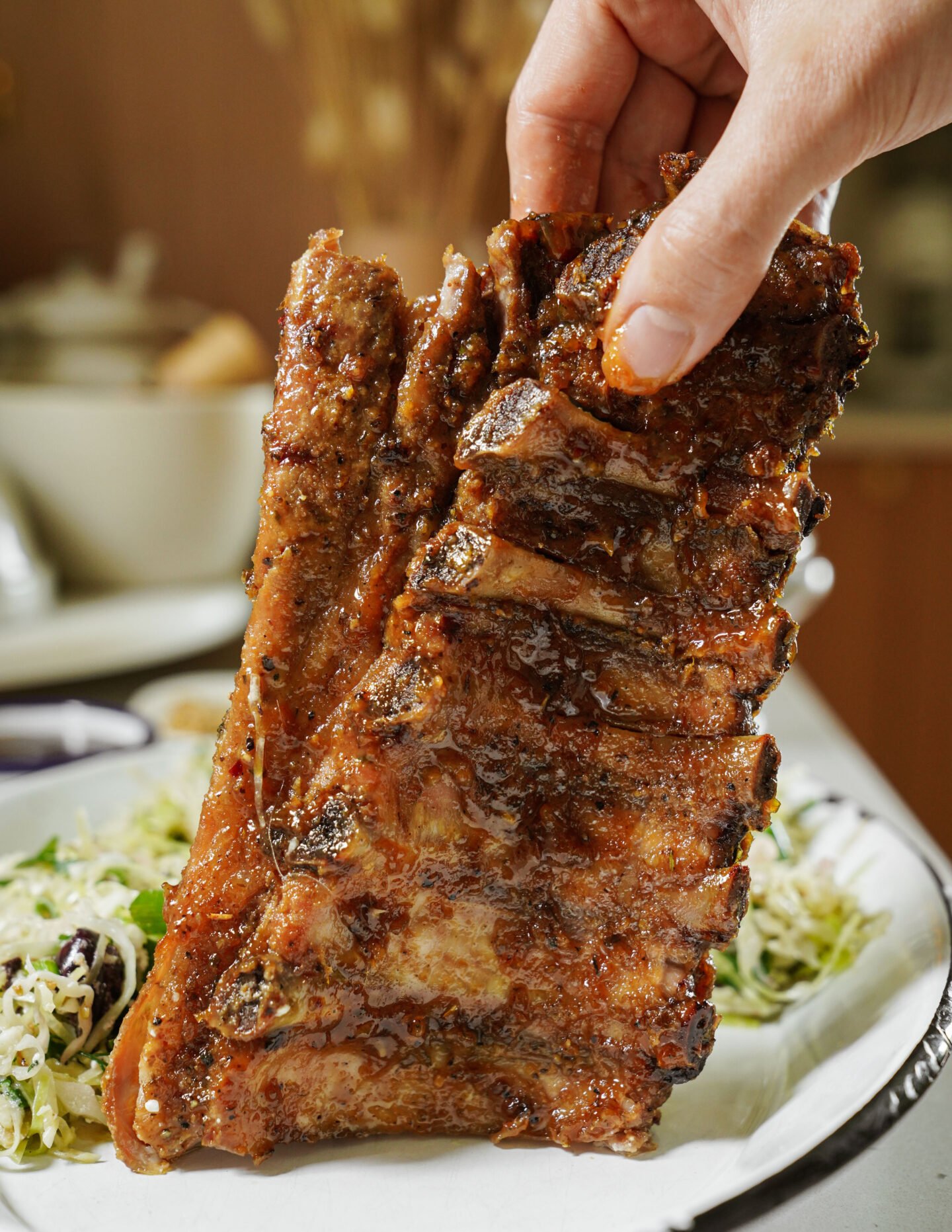 A hand holds up a large, cooked rack of ribs with a glazed, seasoned surface. In the background, there is a plate with salad and blurred tableware.