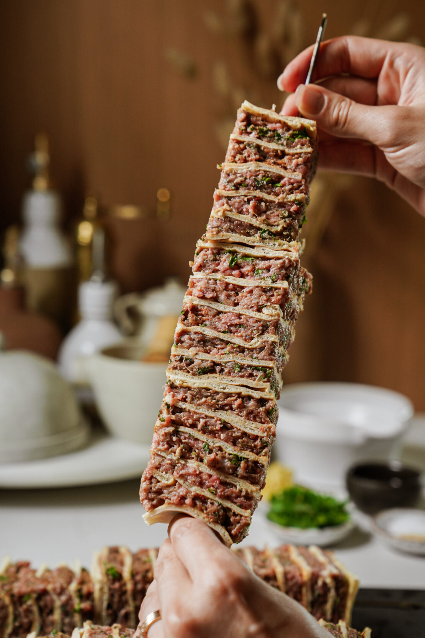 A close-up of hands holding a skewer with multiple layers of sliced meat and herbs, stacked with thin flatbread in between, with a blurred background of dishes and kitchen items.