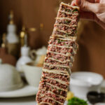 A hand holds up a stack of sliced, layered flatbread filled with ground meat and herbs, with a blurred kitchen background featuring white dishes and utensils.
