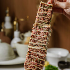 A hand holds up a stack of sliced, layered flatbread filled with ground meat and herbs, with a blurred kitchen background featuring white dishes and utensils.