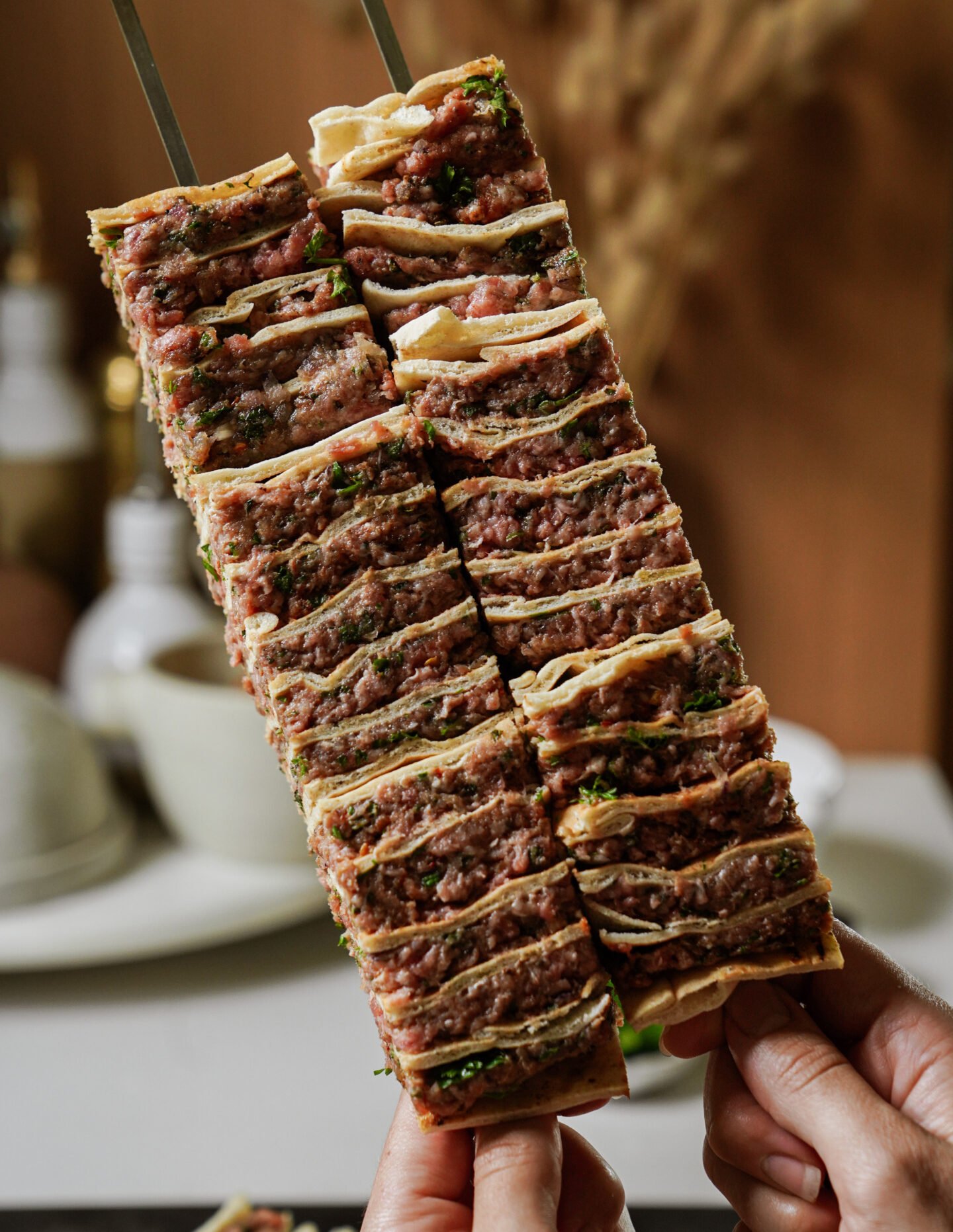 Two hands hold skewers stacked with alternating layers of thin flatbread and seasoned minced meat, ready to be grilled. The background is softly blurred with warm, neutral tones.