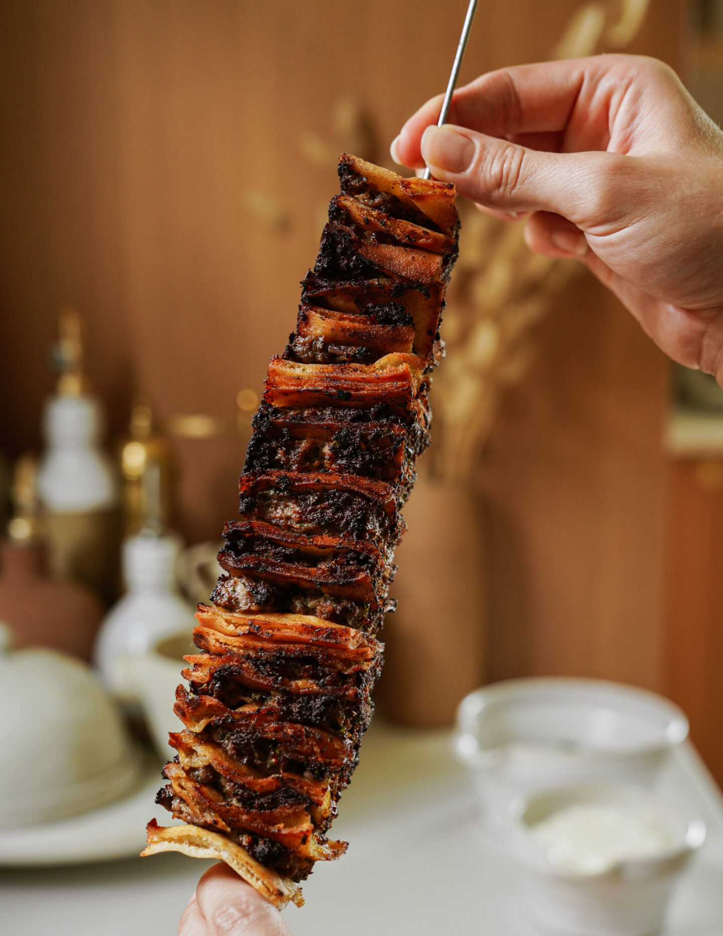 A hand holds a skewer stacked with grilled, seasoned meat slices, slightly charred and crispy, with a blurred background showing kitchen items and a bowl of white sauce.