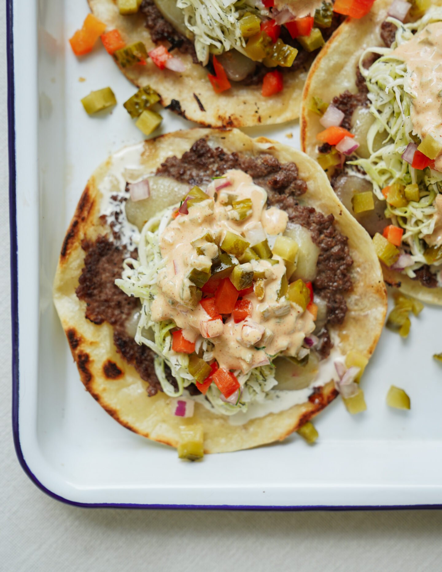 Three open-faced tacos on a tray, topped with ground beef, shredded lettuce, diced pickles, red onions, red bell peppers, and a creamy sauce. The tortillas are slightly toasted.
