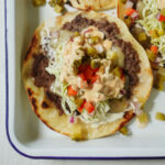 A taco topped with refried beans, shredded lettuce, diced red and green peppers, onions, pickles, and creamy sauce, served on a soft tortilla in a white tray.