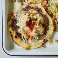 A taco topped with refried beans, shredded lettuce, diced red and green peppers, onions, pickles, and creamy sauce, served on a soft tortilla in a white tray.