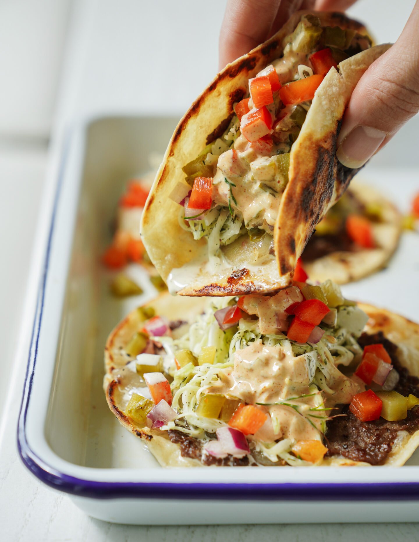 A hand holds a taco filled with shredded vegetables, diced red peppers, creamy sauce, and grilled meat, above a tray with more assembled tacos on a white surface.