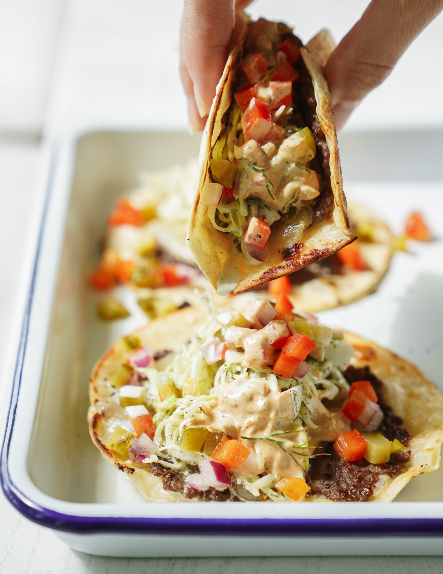 A hand holds a taco filled with ground meat, shredded lettuce, diced red peppers, onions, pickles, and a creamy sauce, above a tray with two more loaded tacos.