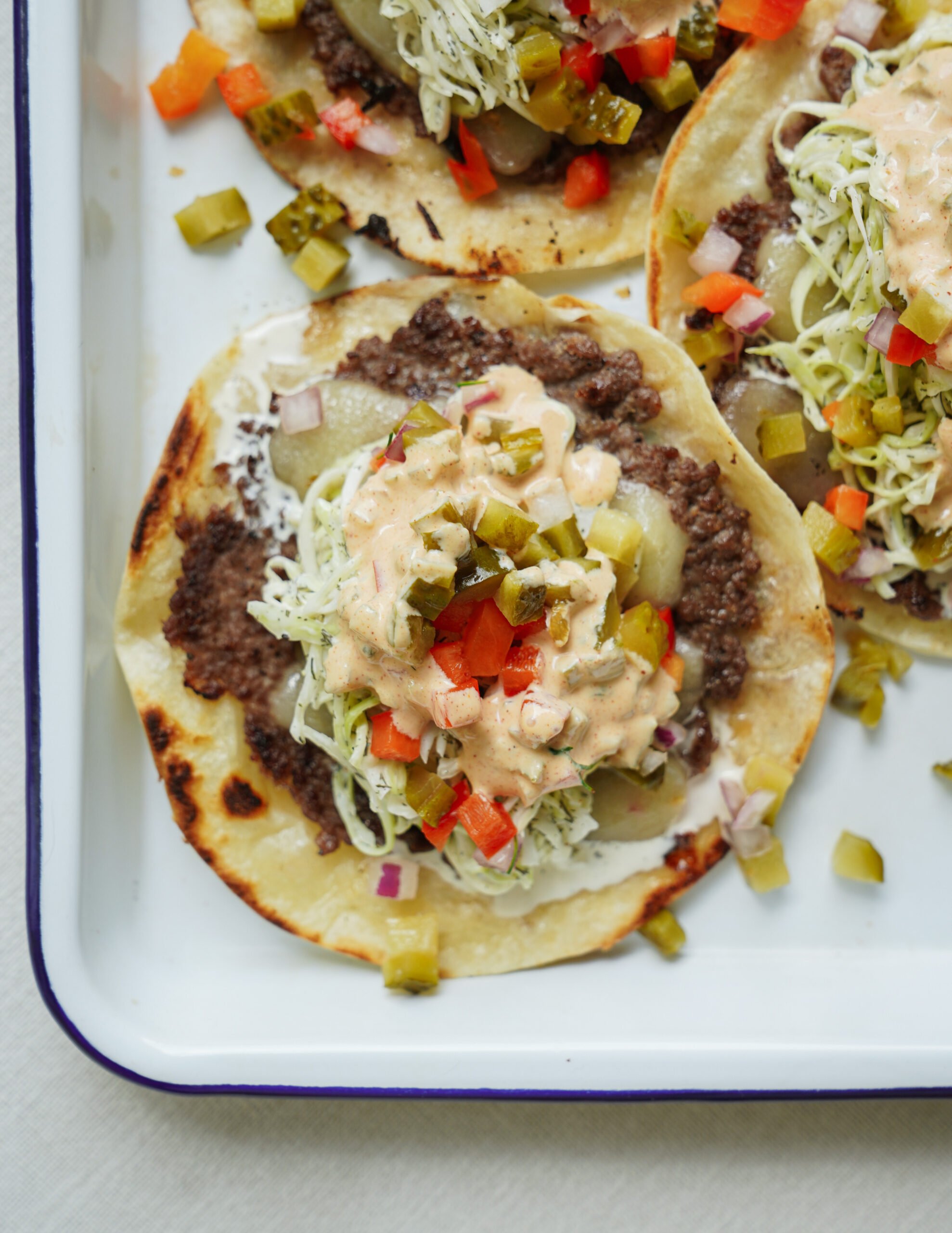 Three open-faced tacos on a tray, topped with ground beef, shredded lettuce, diced pickles, red onions, red bell peppers, and a creamy sauce. The tortillas are slightly toasted.