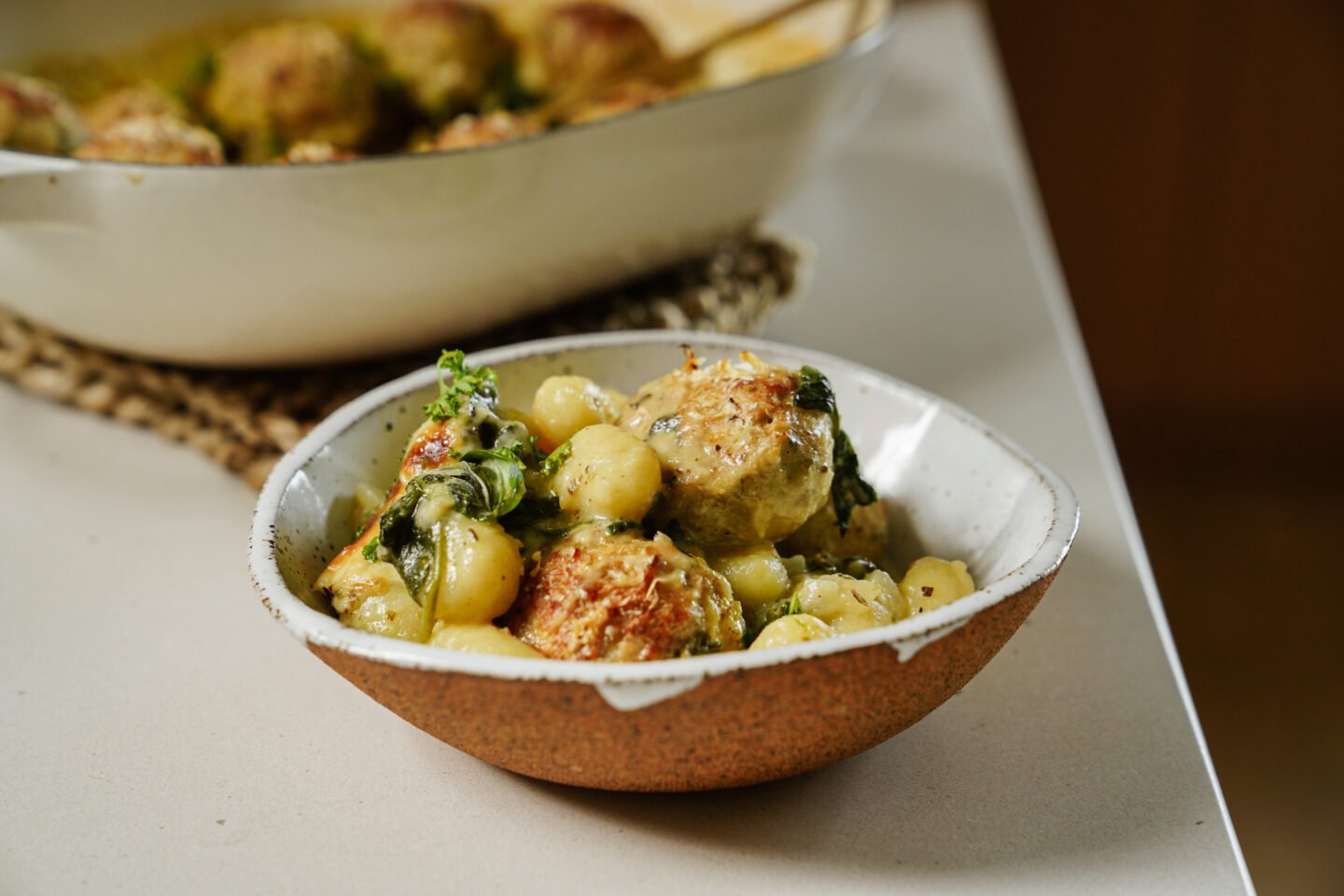 A ceramic bowl filled with gnocchi, spinach, and browned meatballs in a creamy sauce sits on a light countertop. A larger dish of the same meal is visible in the background.