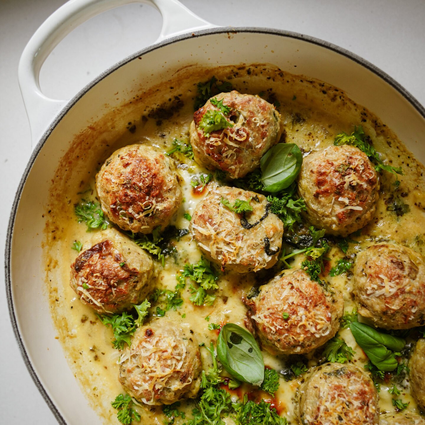 A white baking dish filled with golden-brown meatballs in a creamy sauce, garnished with chopped parsley and fresh basil leaves.