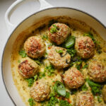 A white baking dish filled with golden-brown meatballs in a creamy sauce, garnished with chopped parsley and fresh basil leaves.