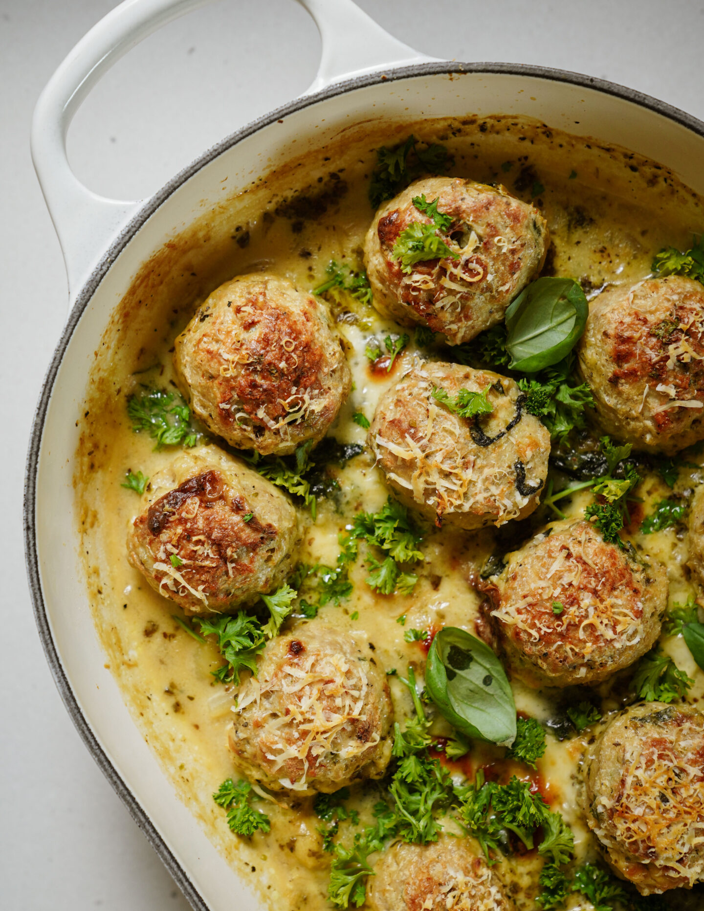 A white casserole dish filled with baked meatballs topped with grated cheese and fresh parsley, sitting in a creamy sauce and garnished with basil leaves.