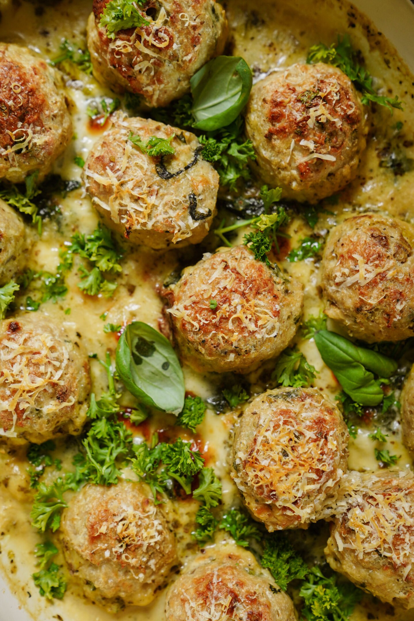 A close-up of golden-brown meatballs topped with grated cheese, fresh parsley, and basil leaves, served in a creamy sauce.