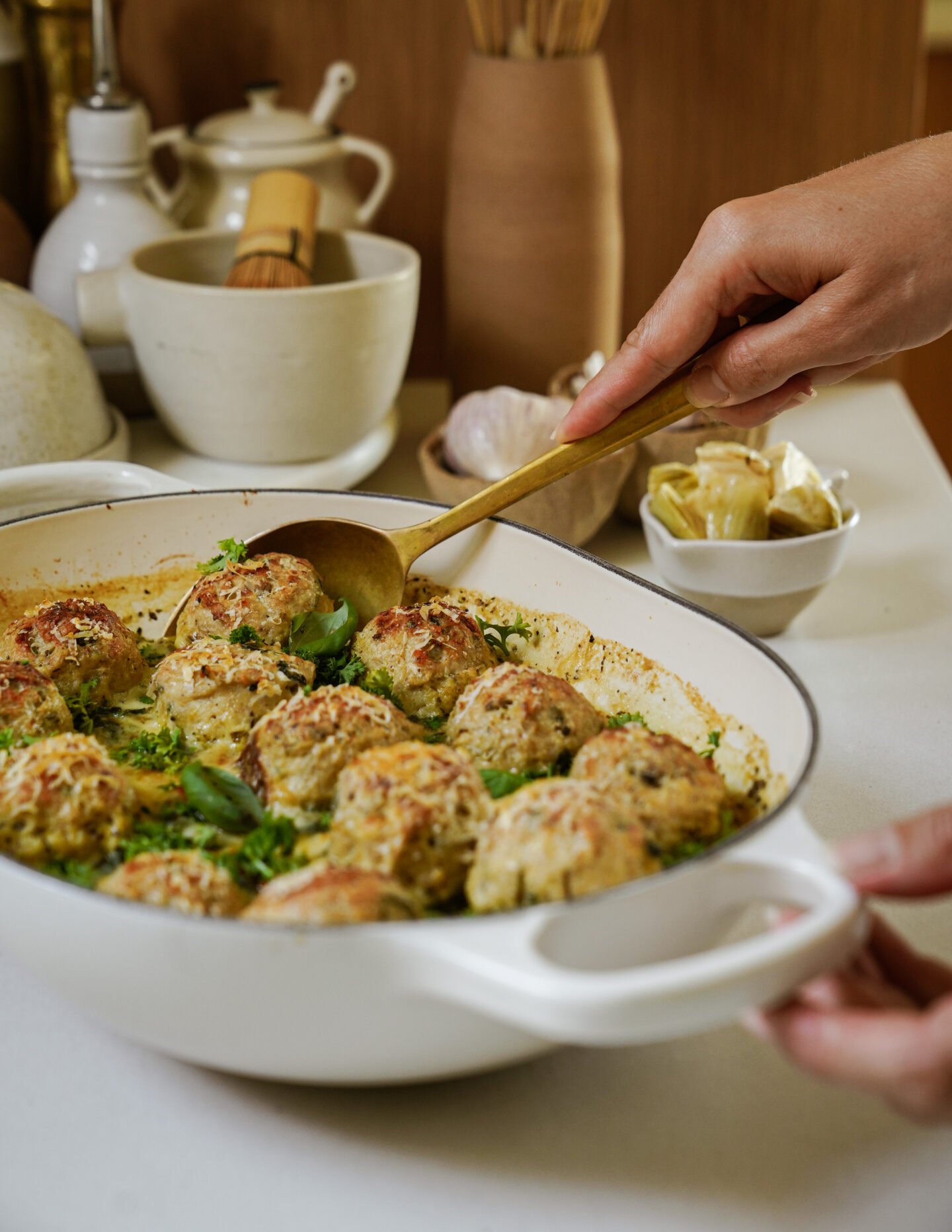 A person serves golden-brown baked meatballs from a white dish with a spoon in a kitchen, with herbs sprinkled on top and kitchen utensils and small dishes in the background.