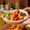 A close-up of hands holding a bowl filled with orzo pasta, chickpeas, roasted cherry tomatoes, chicken pieces, and garnished with fresh basil leaves. The background shows a kitchen setting.