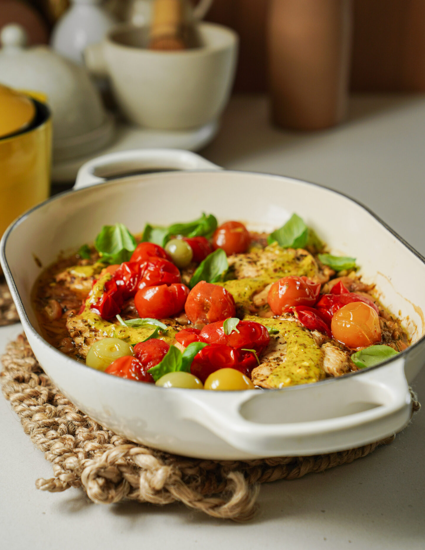 A white baking dish filled with baked chicken breasts, cherry tomatoes, and fresh basil leaves sits on a woven mat on a kitchen counter. The background shows blurred kitchen items.