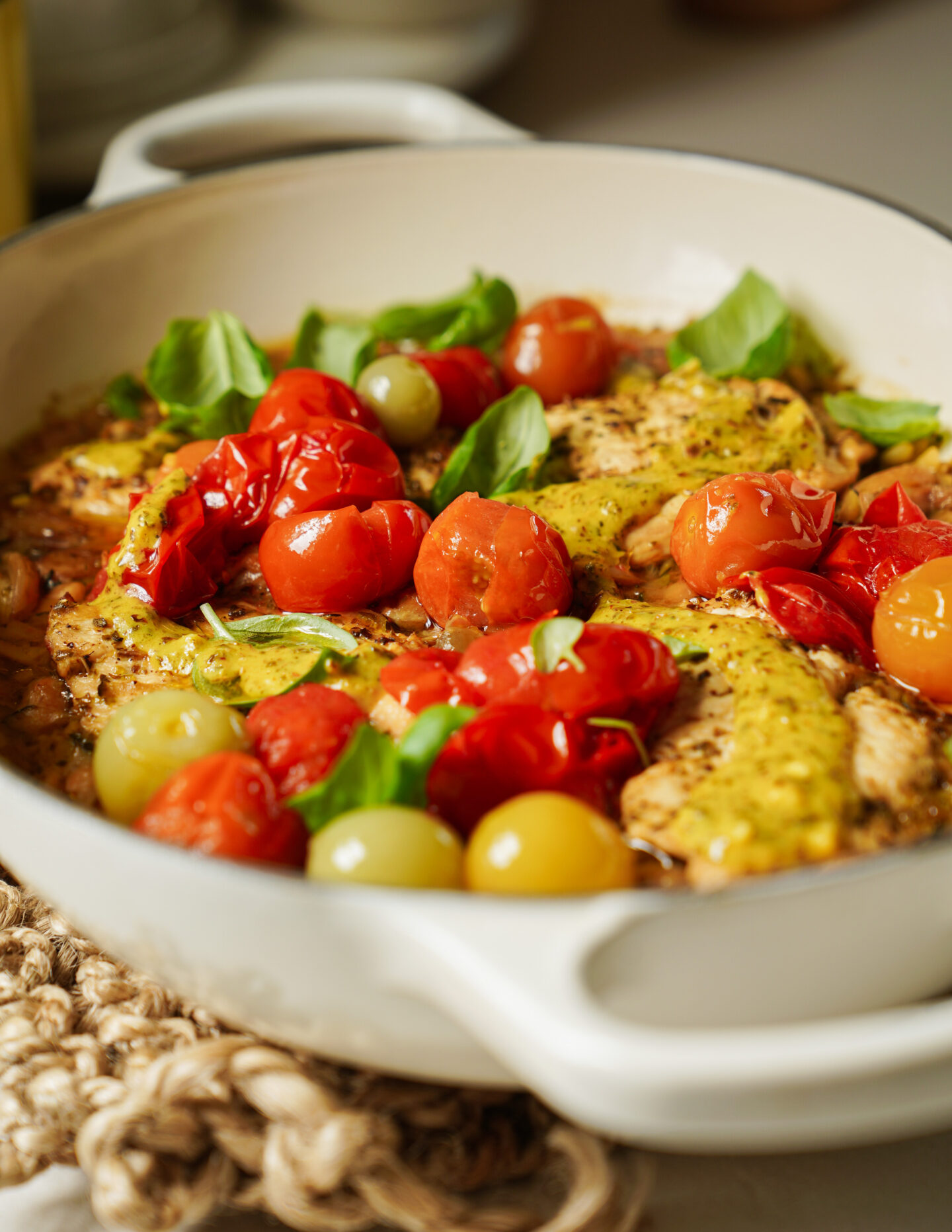 A white dish filled with cooked chicken breast, cherry tomatoes in red and yellow, fresh basil leaves, and a yellow sauce, sitting on a woven mat.