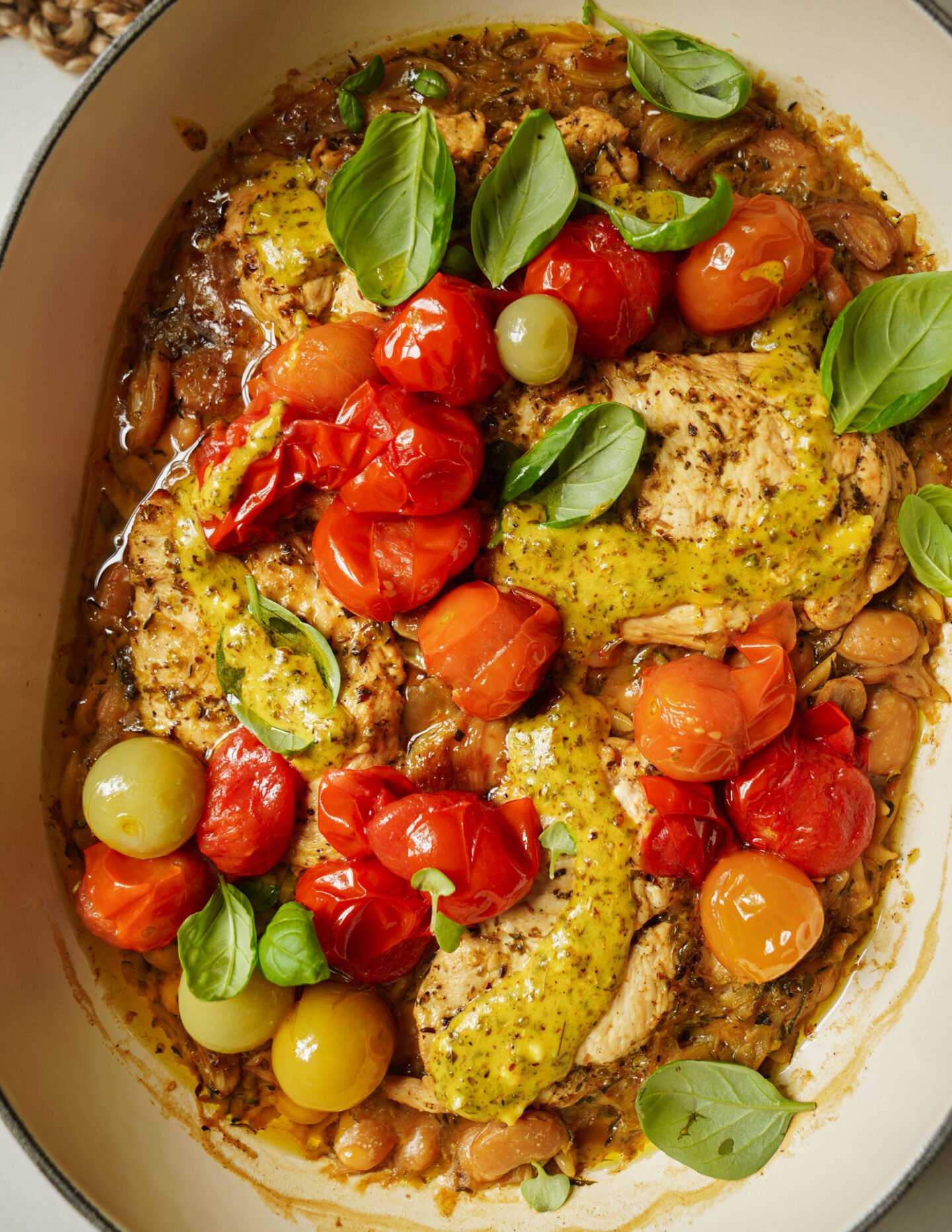 A dish with cooked chicken breasts, roasted cherry tomatoes in red, yellow, and green, fresh basil leaves, and a yellow-green sauce, all in a white casserole dish.