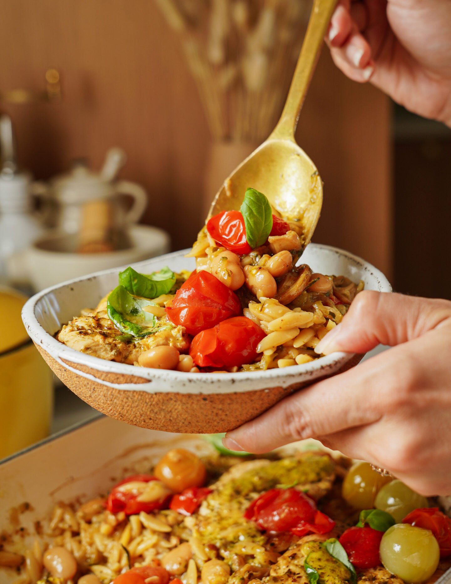 A hand holds a bowl of orzo pasta with cherry tomatoes, white beans, and fresh basil while another hand serves a spoonful from a pot below. The dish is colorful and garnished with herbs.