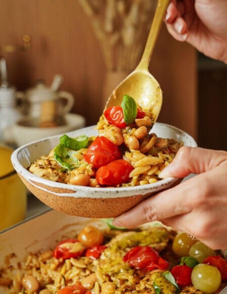 A hand holds a bowl of orzo pasta with cherry tomatoes, white beans, and fresh basil while another hand serves a spoonful from a pot below. The dish is colorful and garnished with herbs.