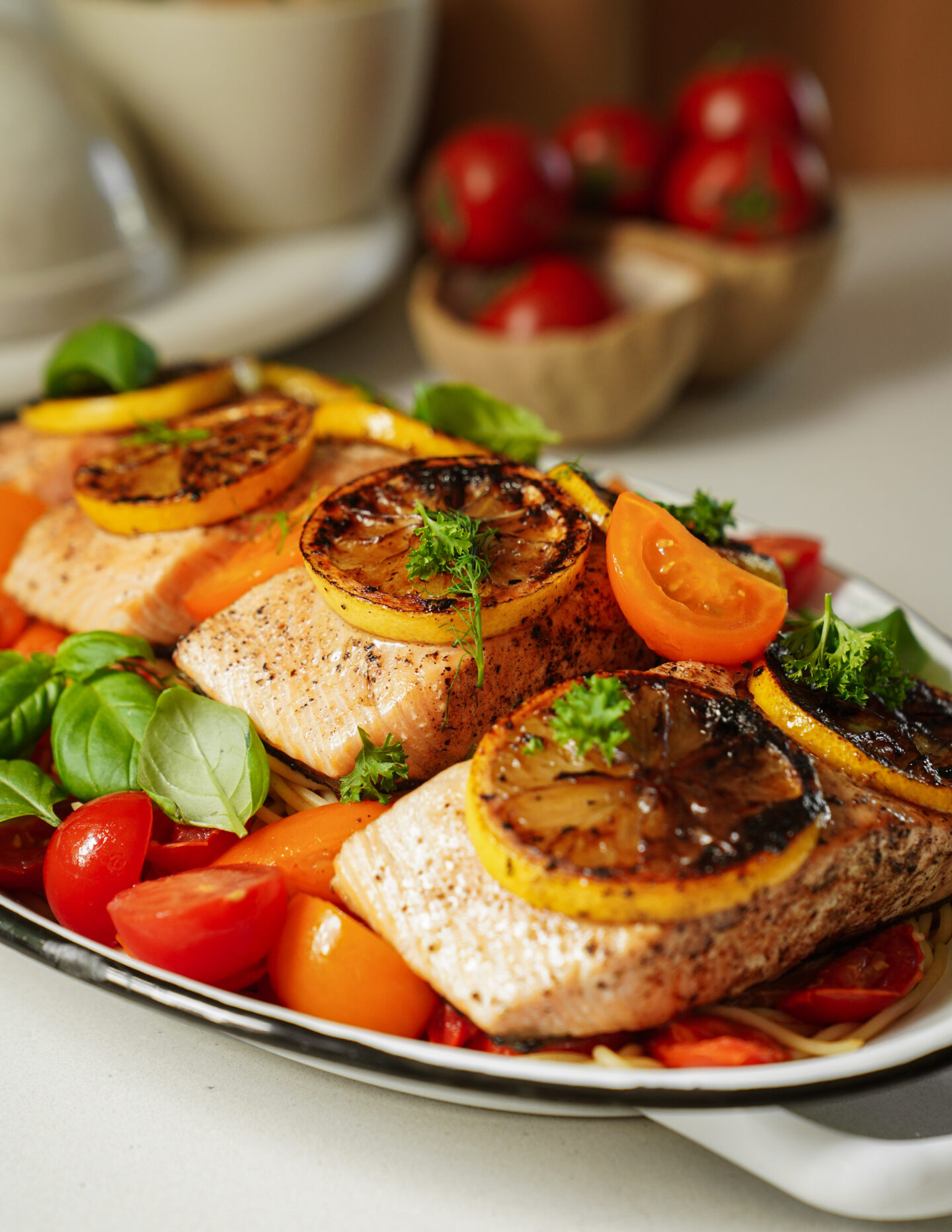 Grilled salmon fillets topped with charred lemon slices, garnished with parsley, and served on a bed of fresh cherry tomatoes and basil leaves on a plate. Eggs and tomatoes are visible in the background.