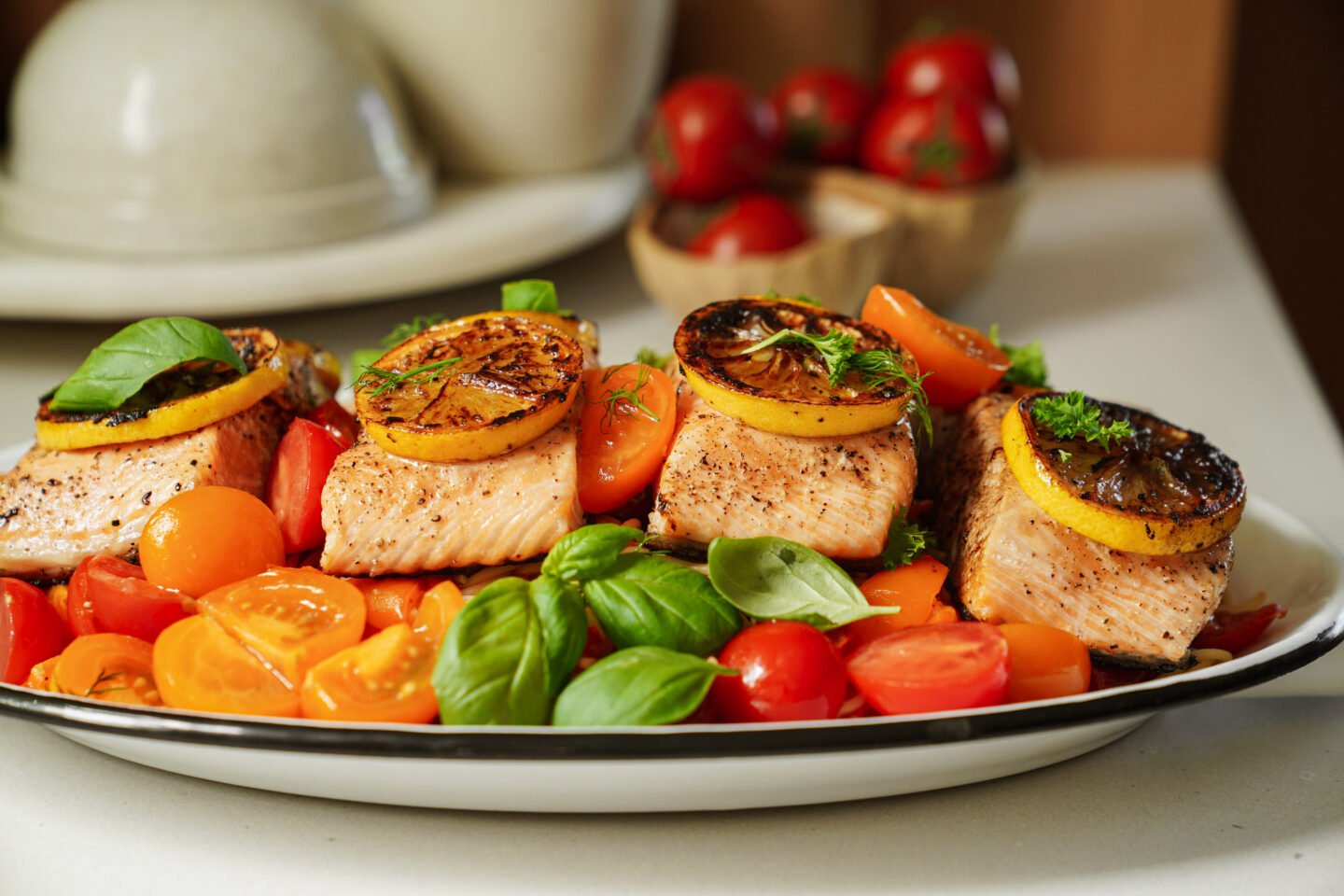 Plated salmon fillets topped with grilled lemon slices, surrounded by halved cherry tomatoes and fresh basil leaves on a white dish. Bowls of tomatoes and plates are in the background.