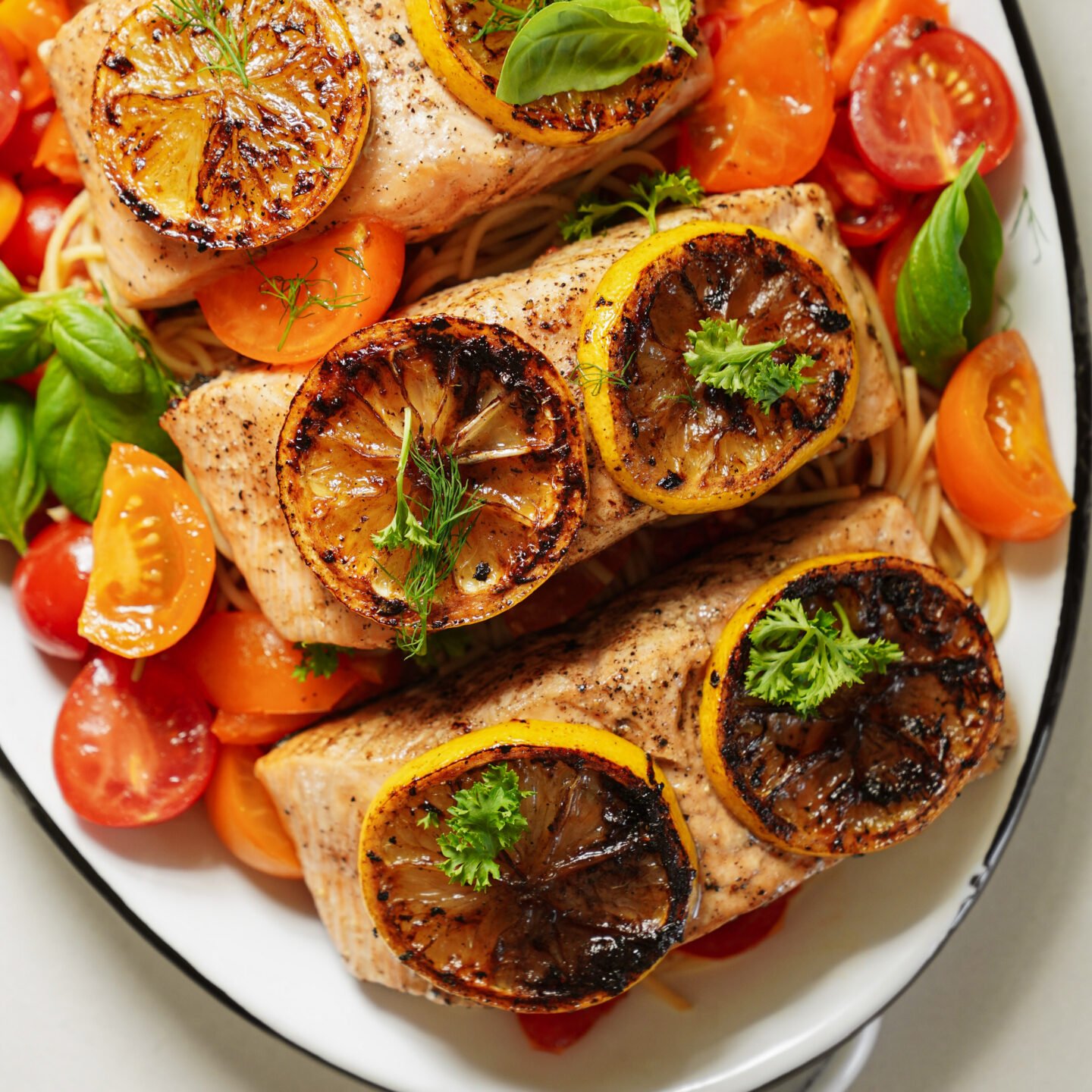 Three pieces of cooked salmon topped with charred lemon slices and herbs sit on a bed of spaghetti with halved cherry tomatoes and fresh basil on a white plate.
