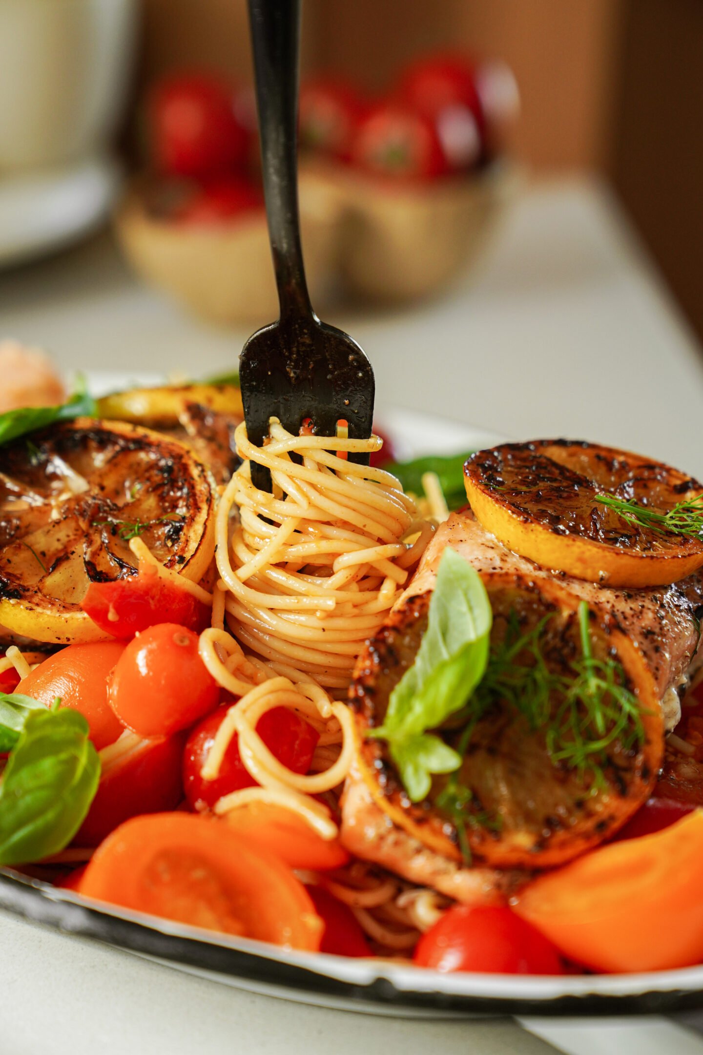 A fork twirls spaghetti on a plate garnished with grilled lemon slices, cherry tomatoes, fresh basil, and herbs. In the background, a bowl of tomatoes is slightly out of focus.