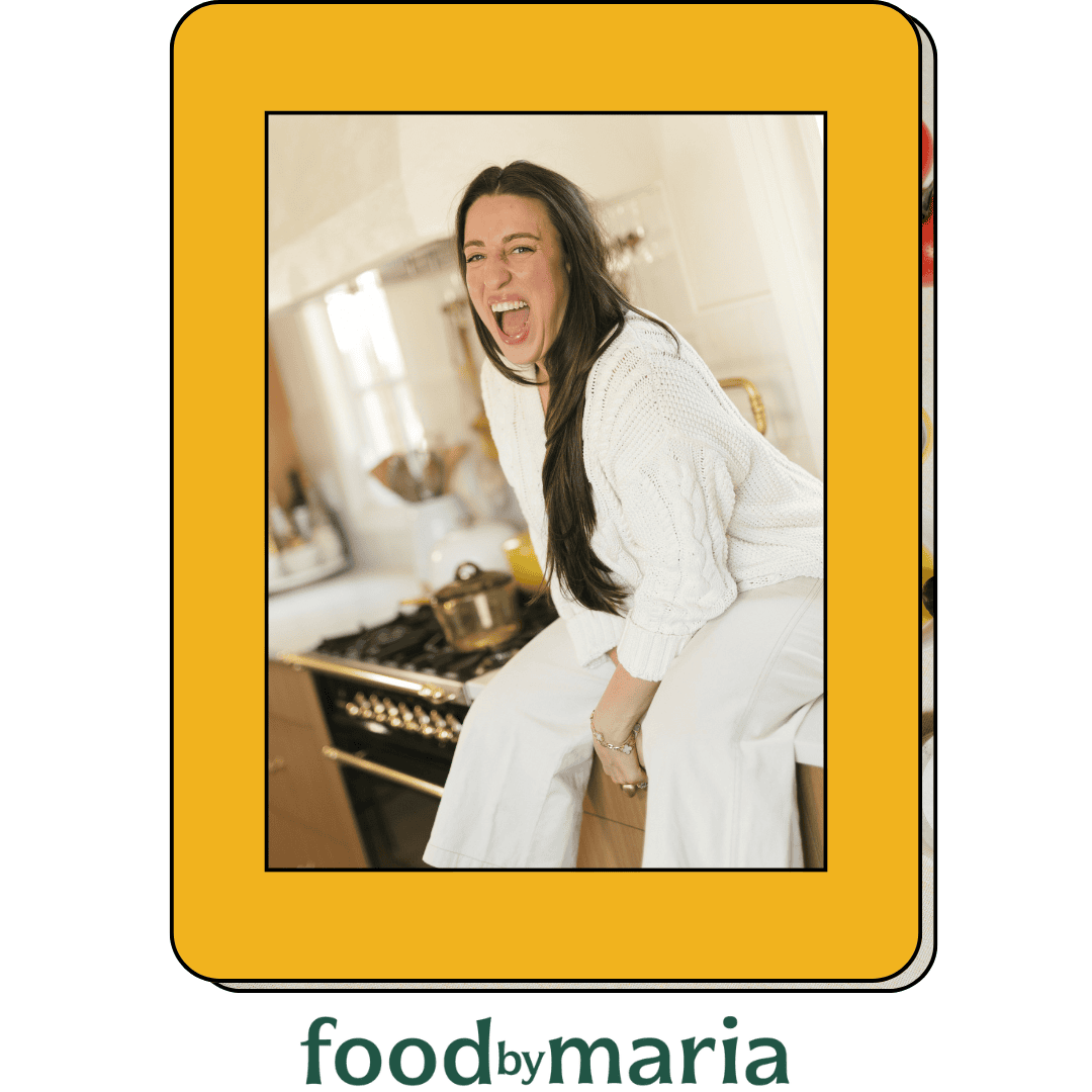 A woman with long dark hair, wearing a white sweater and white pants, sits on a kitchen counter laughing. The image is framed in yellow with the text “food by maria” at the bottom.