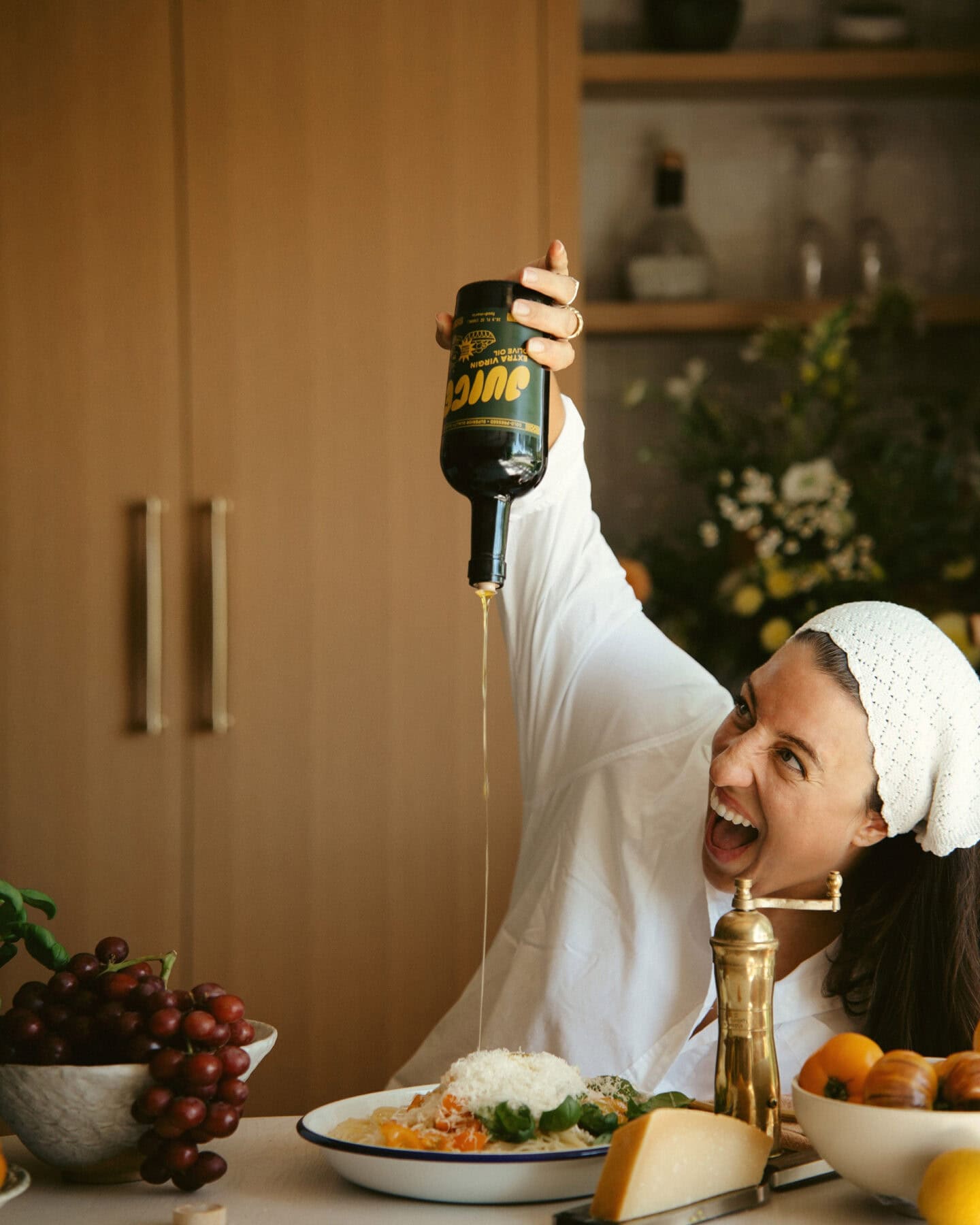 A woman in a white outfit and headband enthusiastically pours olive oil from a large bottle onto a dish of pasta at a table set with grapes, cheese, and a pepper mill. She is smiling widely and leaning forward.