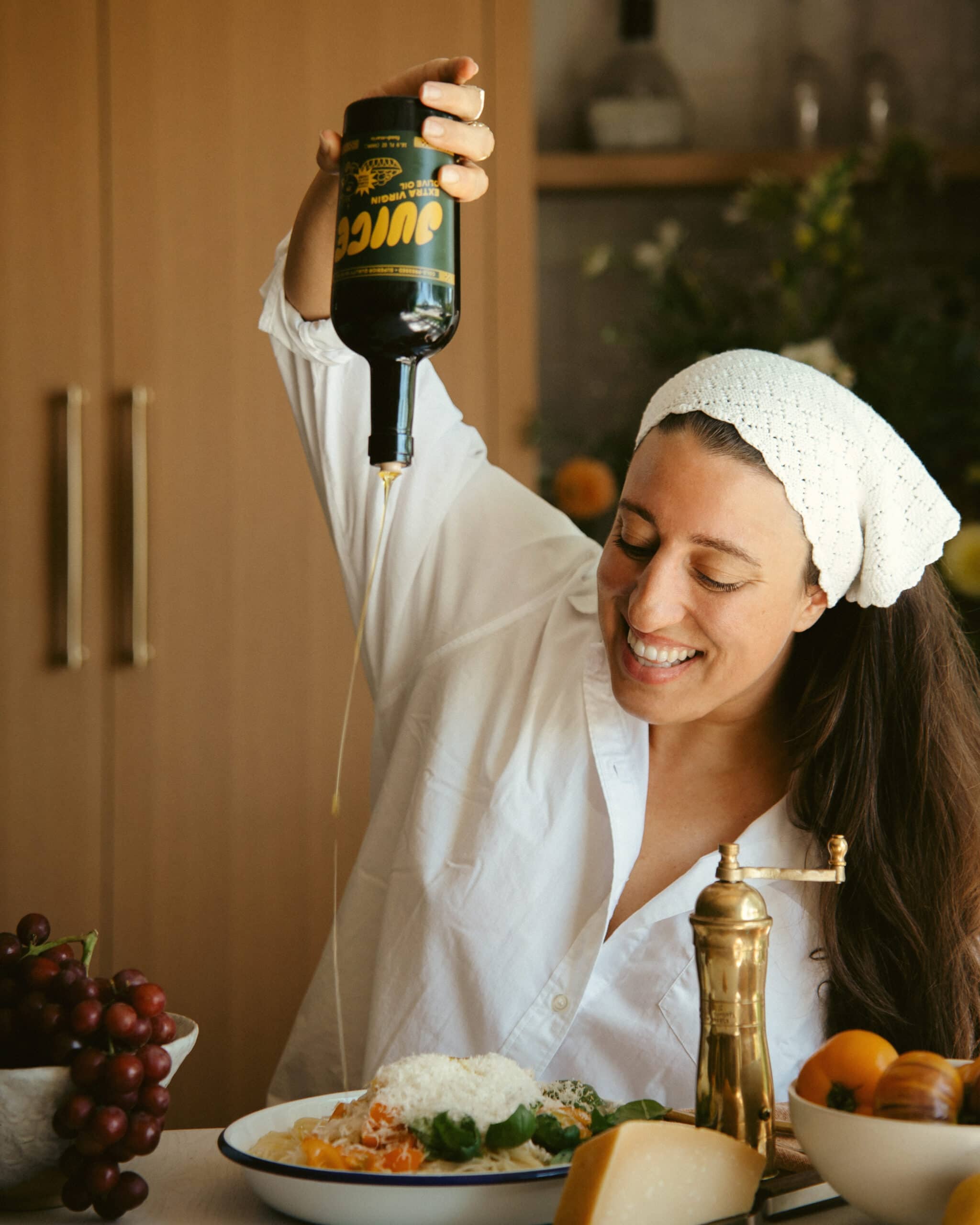 A smiling woman in a white shirt and headscarf pours olive oil from a bottle onto a plate of pasta at a table with grapes, cheese, and fresh produce.