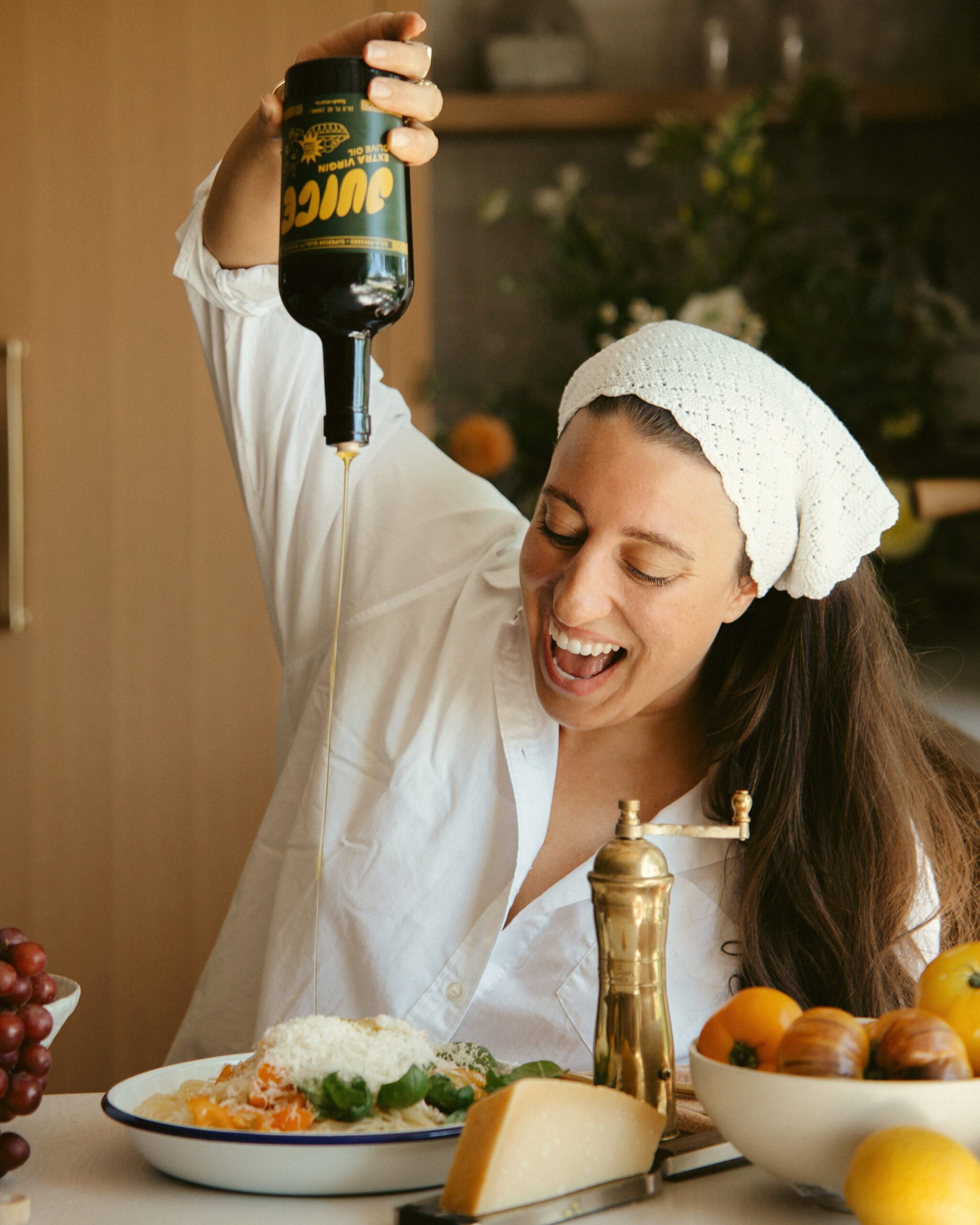 A smiling woman in a white shirt and headscarf enthusiastically pours olive oil onto a plate of food in a kitchen, surrounded by grapes, oranges, cheese, and a pepper grinder.