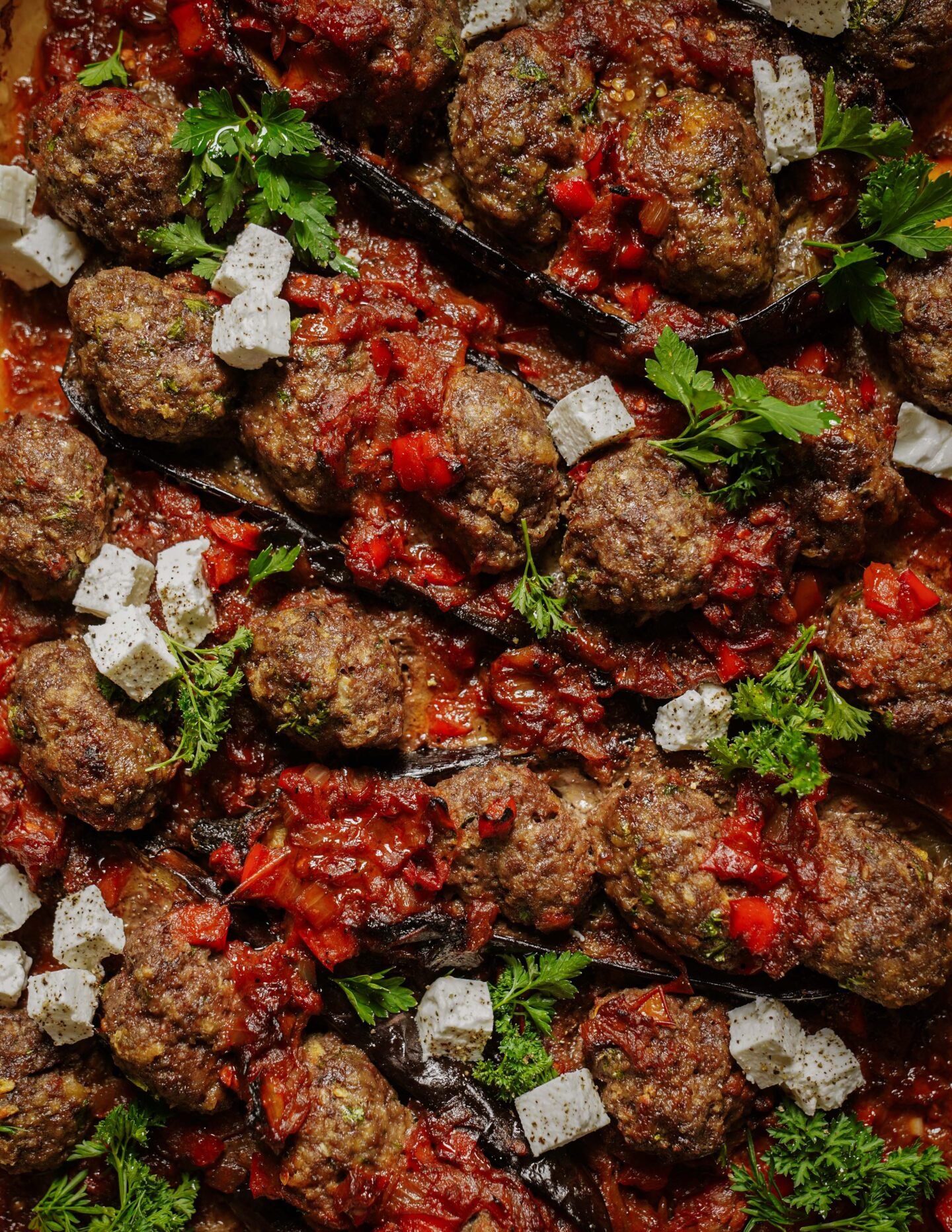 Close-up of baked meatballs with chunky tomato sauce, garnished with fresh parsley and topped with small cubes of white cheese. The dish is colorful with visible herbs and diced red peppers.